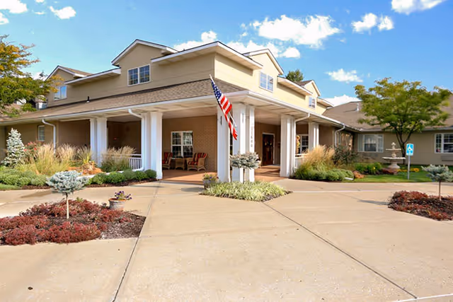 Exterior view of a single-story senior living facility building with a covered entrance supported by white columns. There is an American flag displayed near the entrance, landscaped bushes and small trees around the driveway, and a clear blue sky with some clouds above.