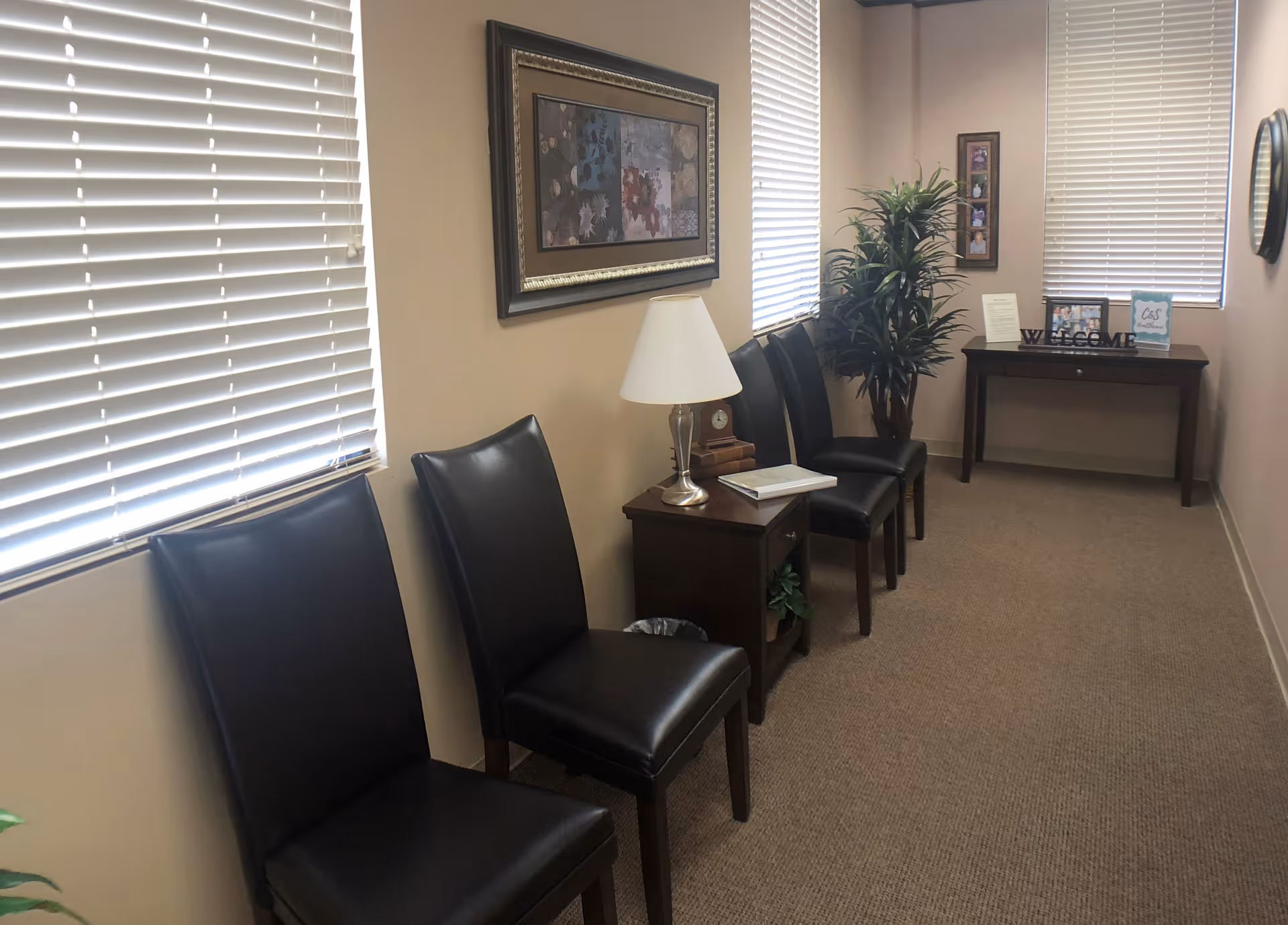 Waiting area with black leather chairs, blinds, a plant, and a welcome desk.