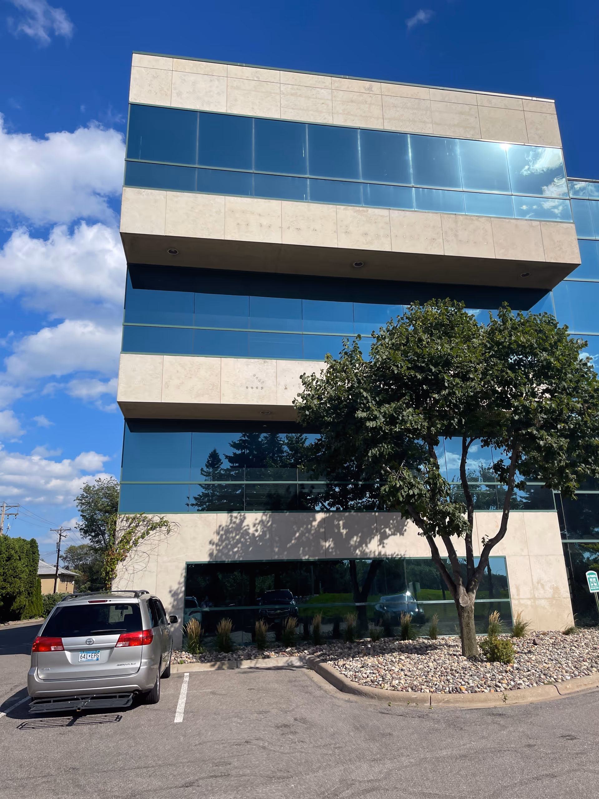 Modern glass-front building with beige stone accents, landscaping, and a parked SUV.