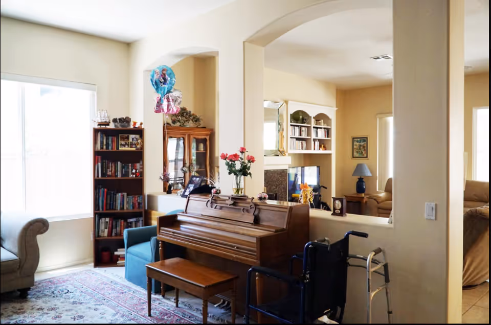 Cozy living room area with a piano, bookshelves, flowers, and a walker near a window.