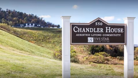 A freestanding sign reading "Chandler House Assisted Living Community" stands on a grassy hillside with buildings visible in the distance.