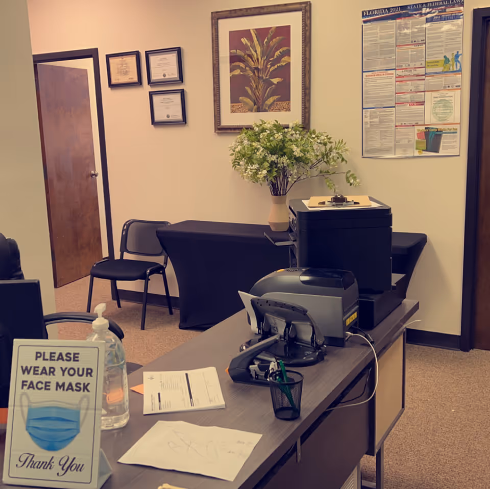 Indoor reception area with desk, printer, mask sign, plant, and wall artwork.
