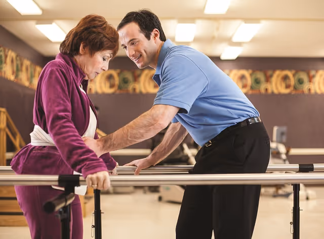 A middle-aged male therapist assists an elderly woman in a purple tracksuit as she uses parallel bars for physical therapy in a rehabilitation room.