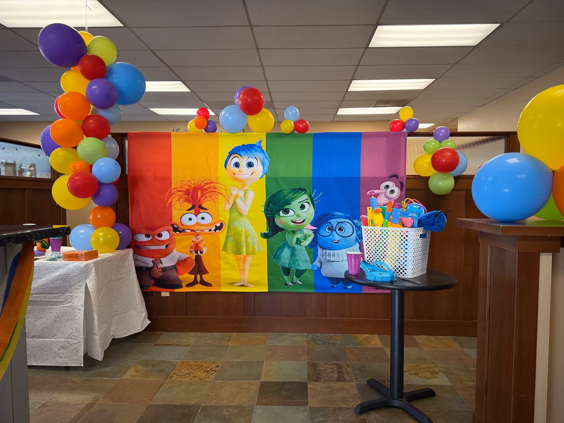 Colorful balloon arch and rainbow cartoon backdrop decorate an interior room at a senior home.