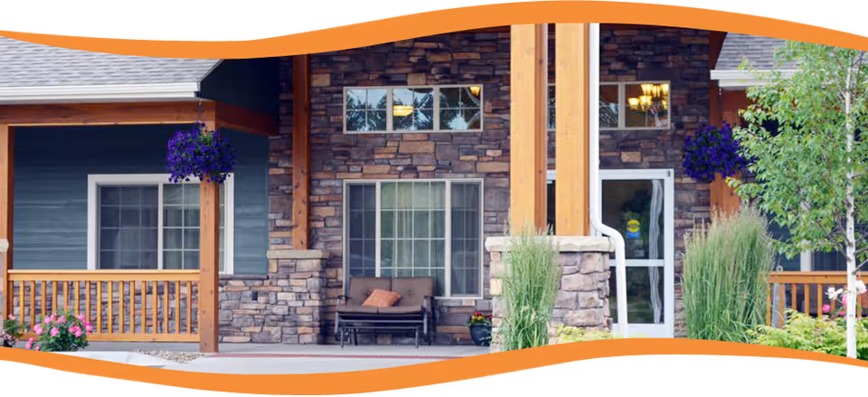 Stone-faced assisted living building entrance with a covered porch, bench seating, hanging flower baskets and landscaped plants.