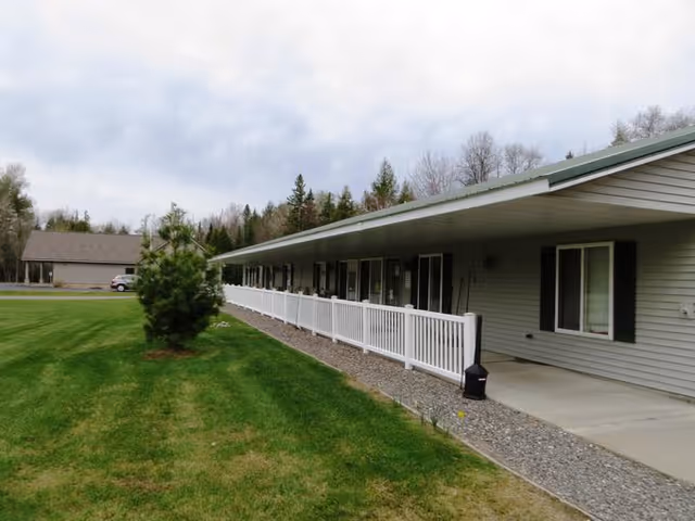 Exterior view of a single-story assisted living facility building with gray siding, a white railing along a covered walkway, and a well-maintained green lawn with a small tree. Another building and a parked vehicle are visible in the background under a cloudy sky.