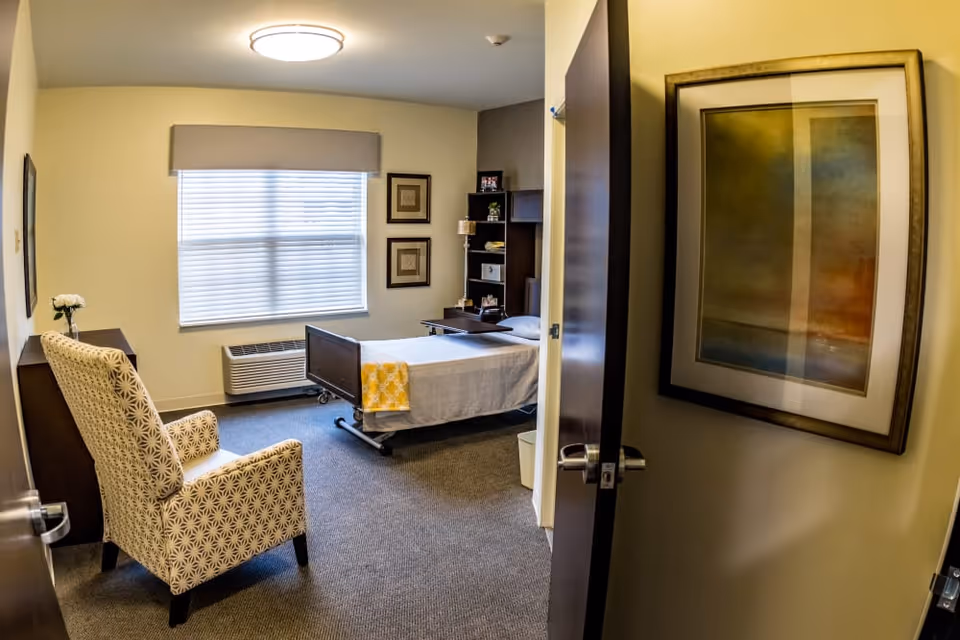 A furnished single bedroom with a bed, armchair, window and framed artwork seen through an open door.
