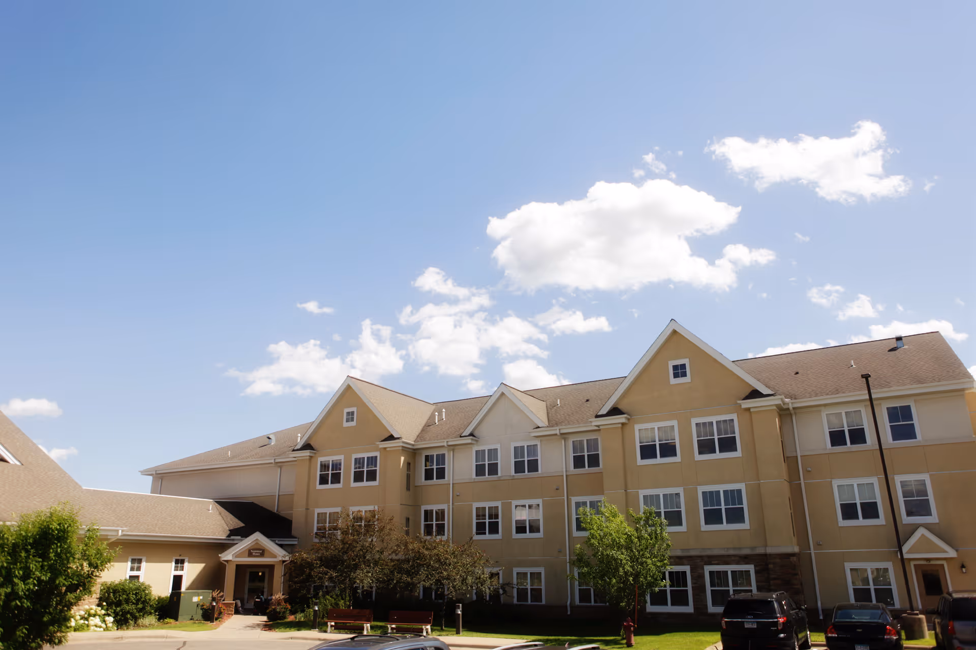 Exterior view of a three-story senior living facility building under a blue sky with scattered clouds. The building has beige walls, multiple windows, and a peaked roof. There are some trees, bushes, benches, and parked cars in front of the building.