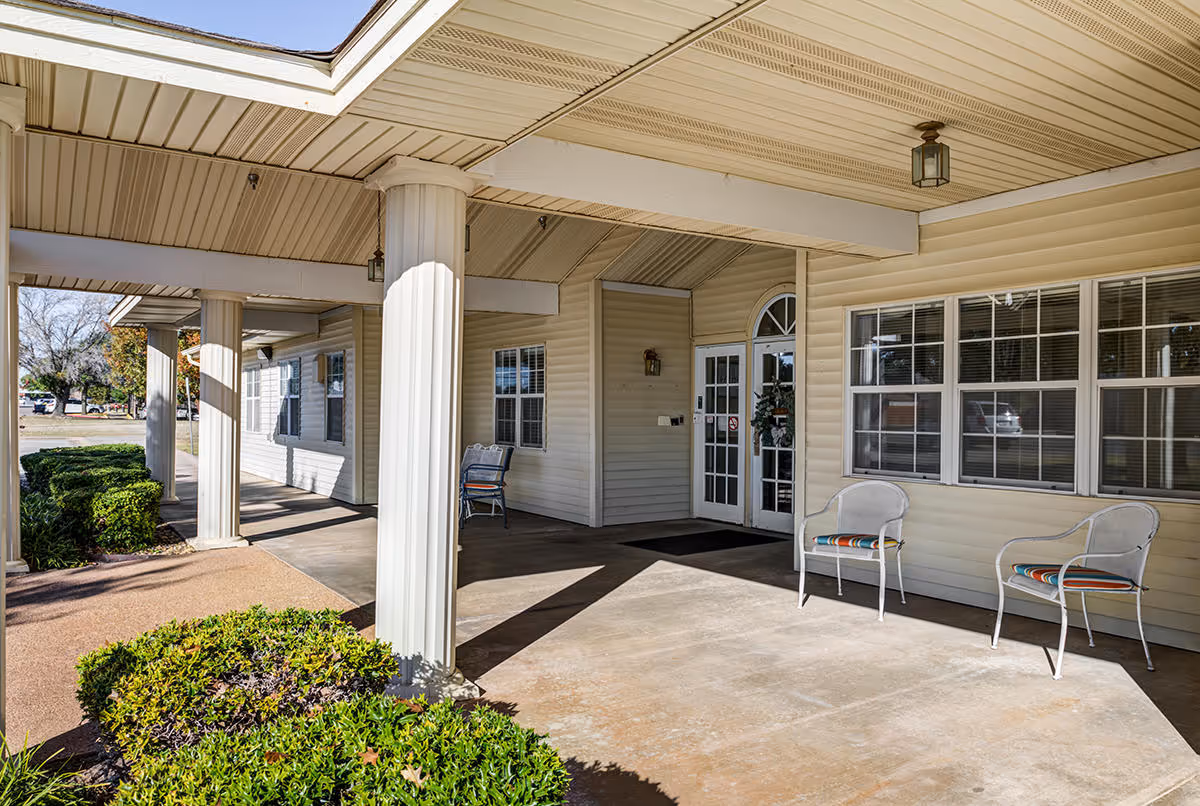 Covered entrance area of a building with white columns and beige siding. There are two white chairs with colorful cushions placed near a window, and a glass door with a wreath hanging on it. Green bushes line the walkway leading to the entrance.
