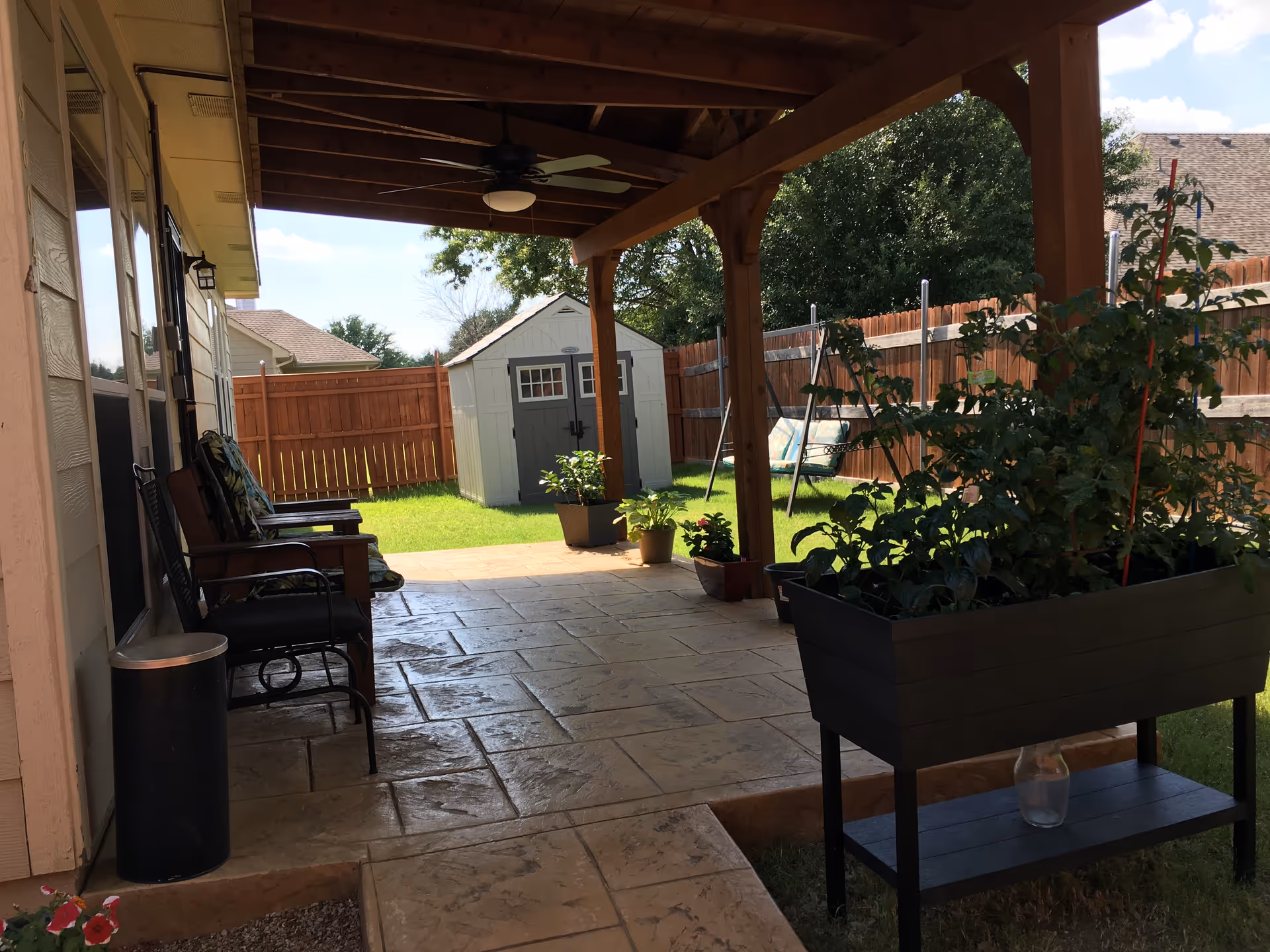 Covered patio with seating, potted plants, a lawn, and a wooden fence.