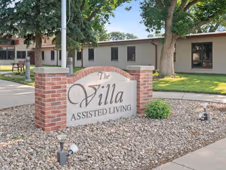 Exterior view of a single-story assisted living facility with a brick sign and landscaping.