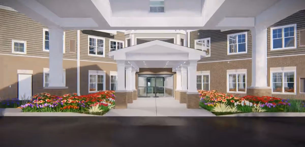Entrance of a senior living facility with a covered driveway, surrounded by flower beds with colorful flowers and a multi-story building with many windows in the background.