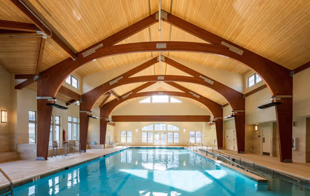 Indoor swimming pool with clear blue water under a high wooden ceiling with large wooden beams. The pool area is well-lit with natural light coming through multiple windows and glass doors at the far end. There are handrails along the pool edges and seating areas on the left side.