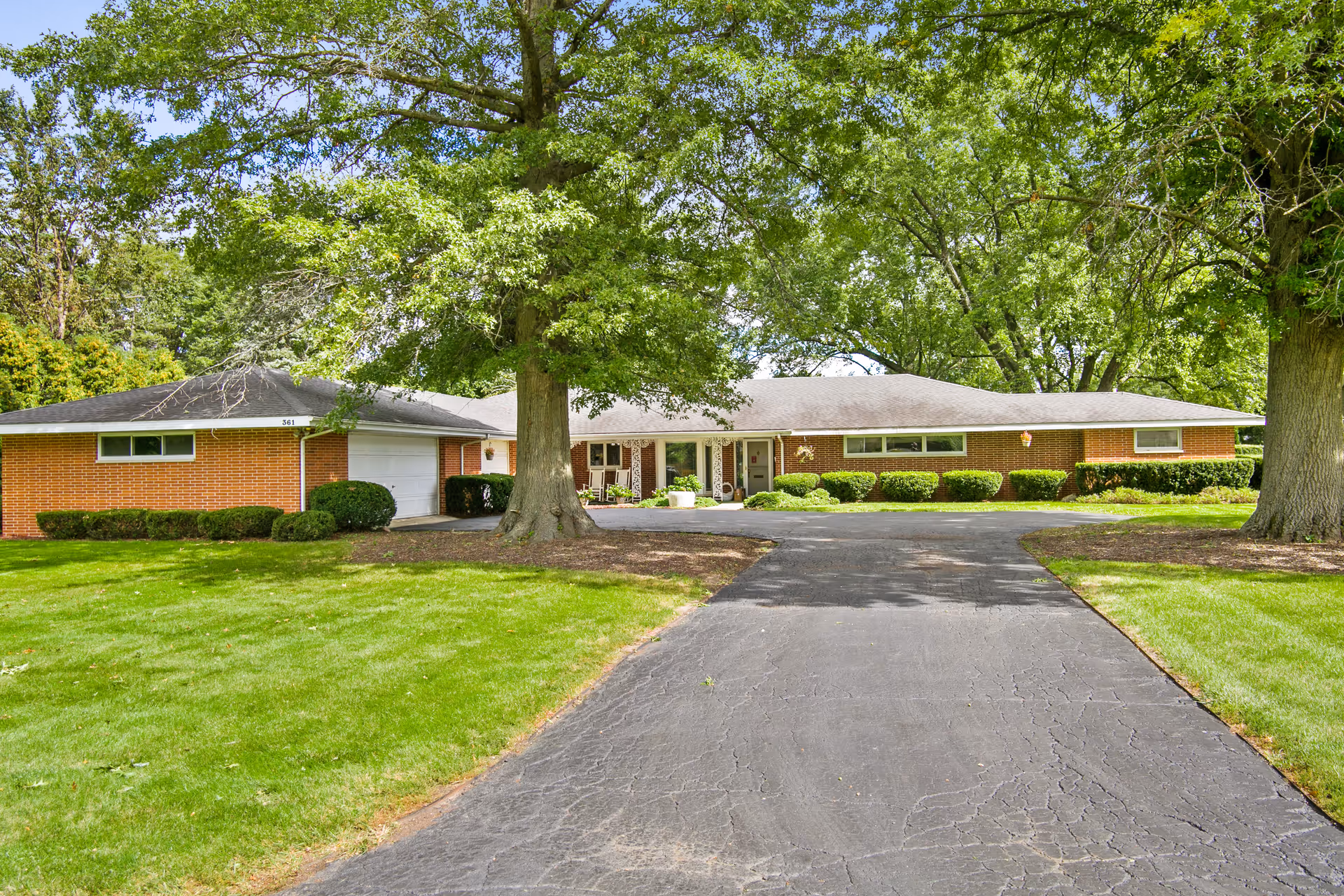 A single-story brick building with a large driveway and well-maintained green lawn. There are two large trees in the front yard providing shade. The building has a garage on the left side and a covered entrance with chairs near the center. The sky is clear and the surrounding area is lush with greenery.