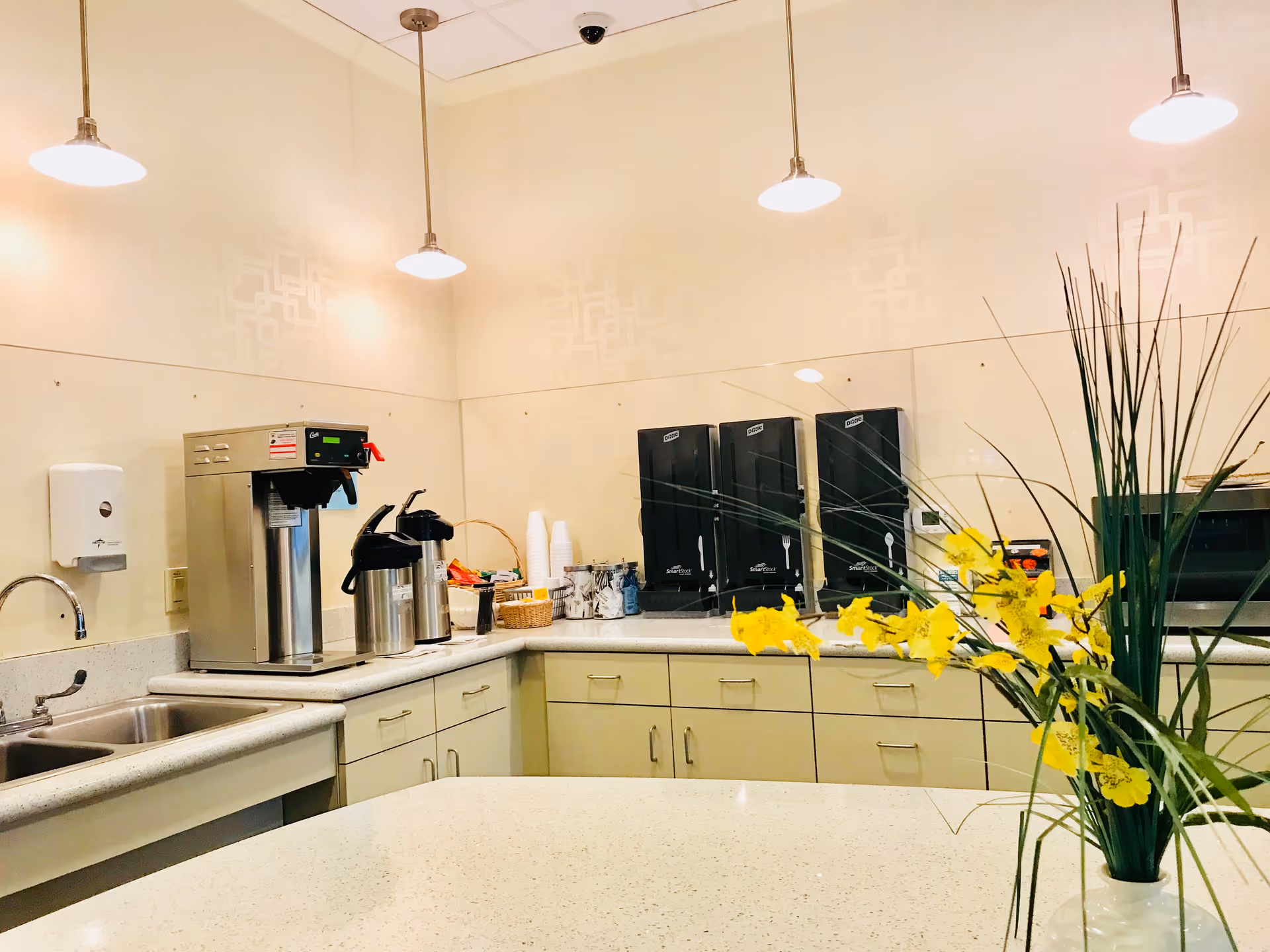 Cream-colored kitchen with coffee station, cup dispensers, microwaves, and yellow flower arrangement on counter.