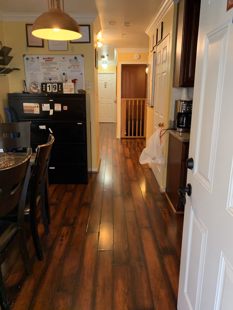 View down a polished wood-floored interior hallway with dining chairs, a black cabinet, and doors leading to rooms.