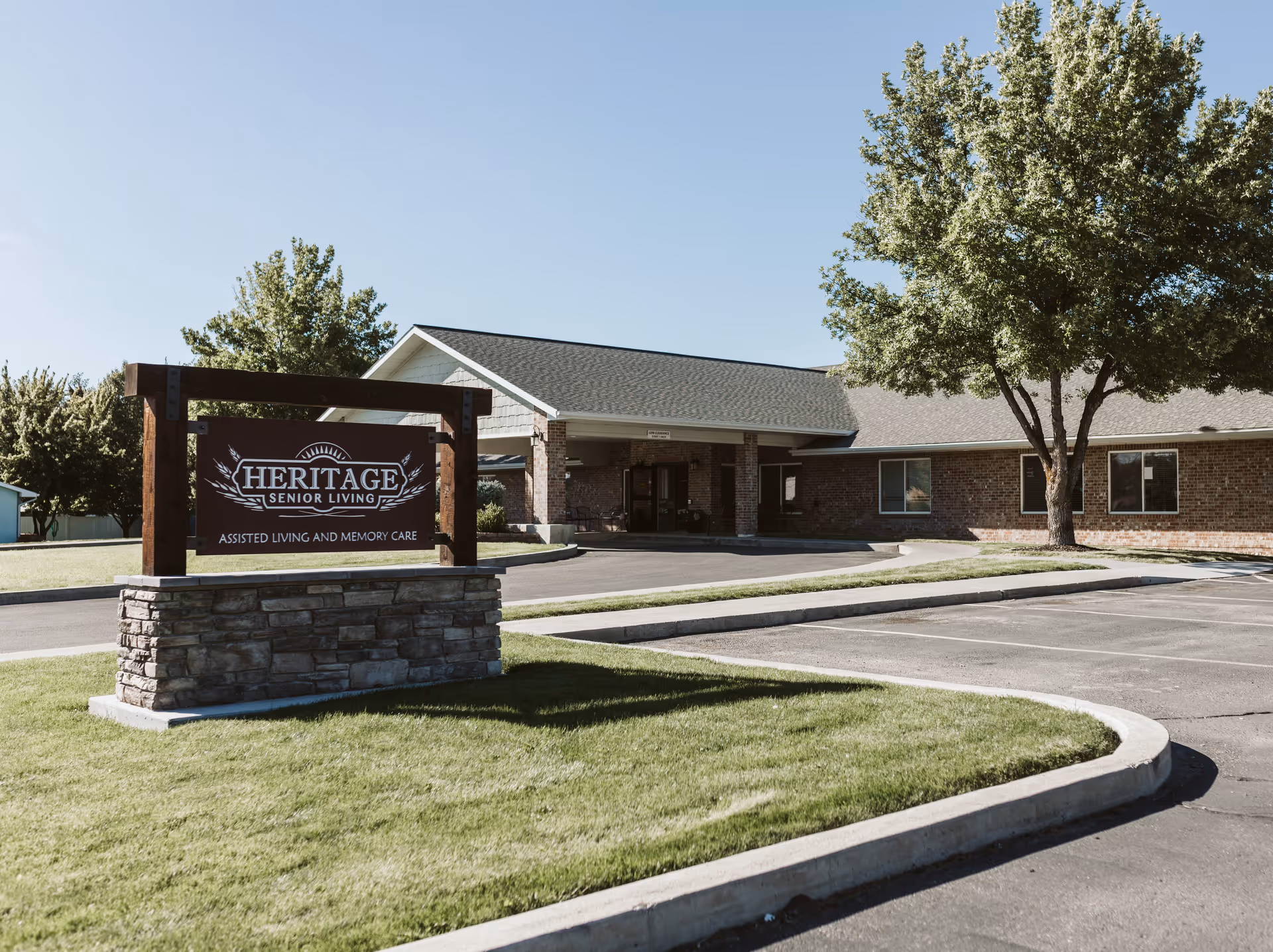 Exterior view of Heritage Senior Living facility showing a single-story brick building with a covered entrance, a large tree, and a sign in the foreground that reads 'Heritage Senior Living Assisted Living and Memory Care'. The area includes a parking lot and well-maintained grassy landscaping under a clear blue sky.
