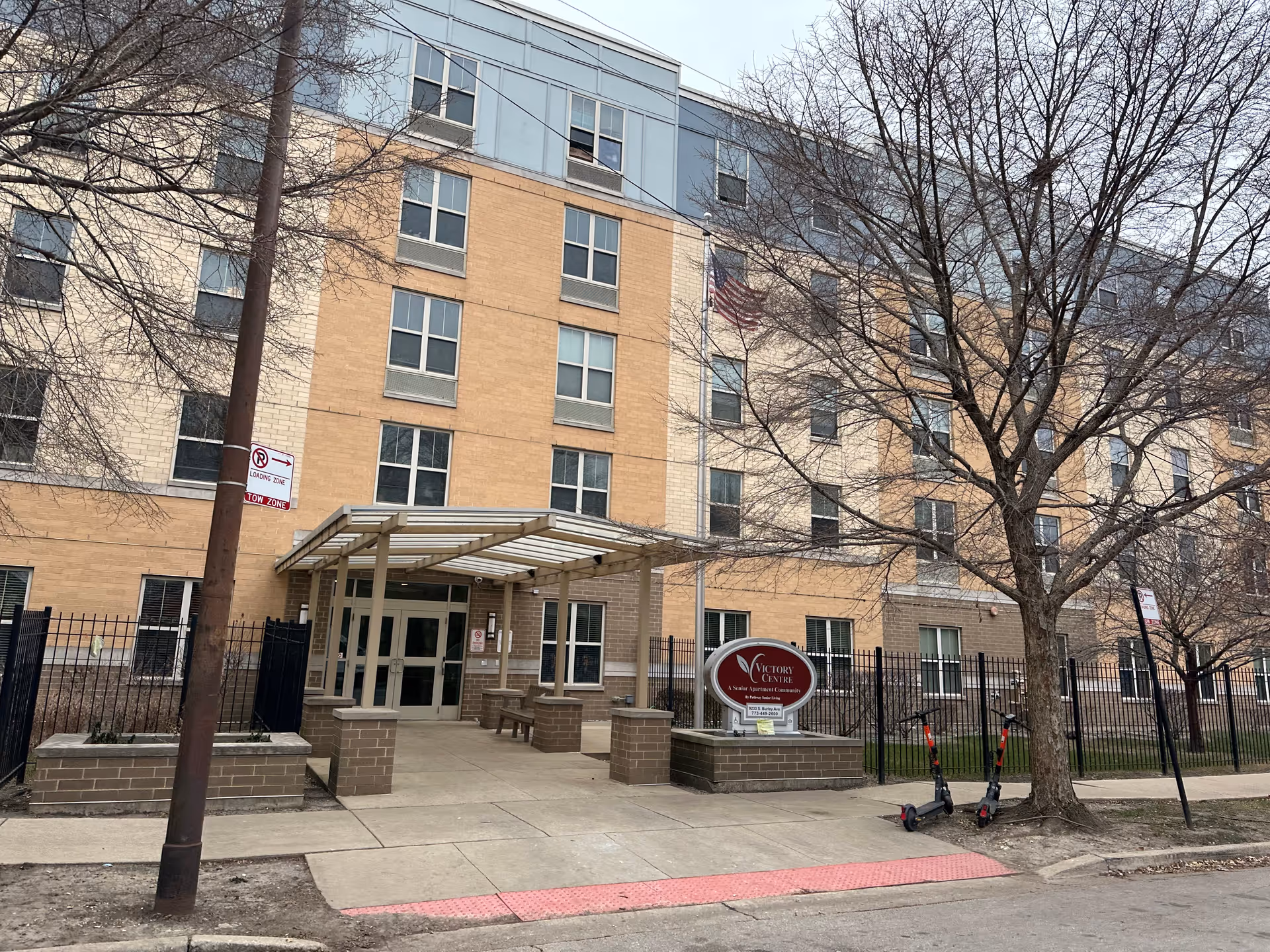 Covered front entrance of a multi-story brick-and-tan senior living building with a 'Victory Centre' sign, flagpole, and parked electric scooters on the sidewalk.
