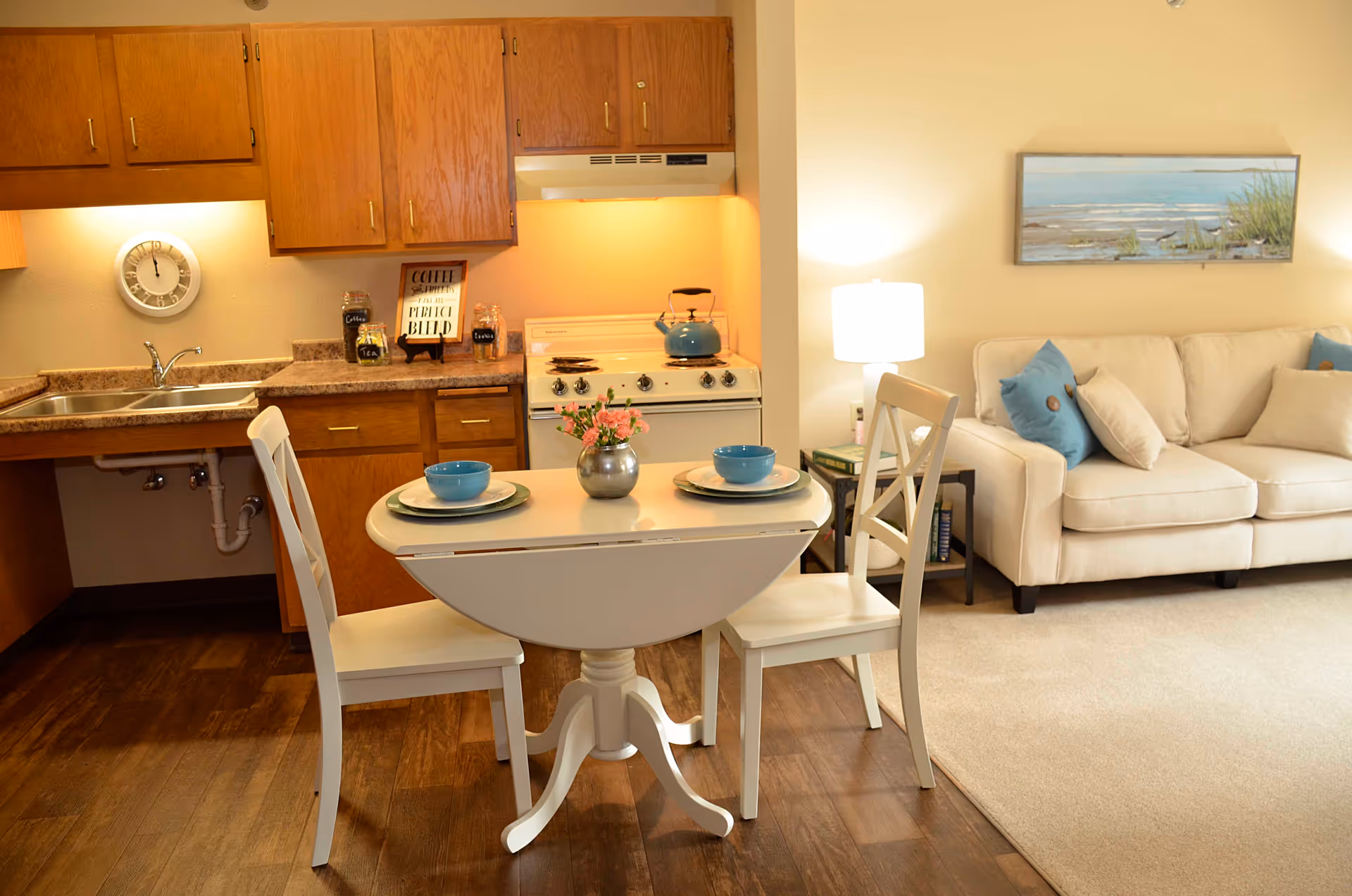 A cozy living space featuring a small kitchen with wooden cabinets, a white stove with a blue kettle, and a double sink. In front of the kitchen is a small white dining table set for two with blue bowls and plates, and a small vase with pink flowers. To the right, there is a beige sofa with blue and beige cushions, a side table with books, a lamp, and a framed beach-themed painting on the wall.