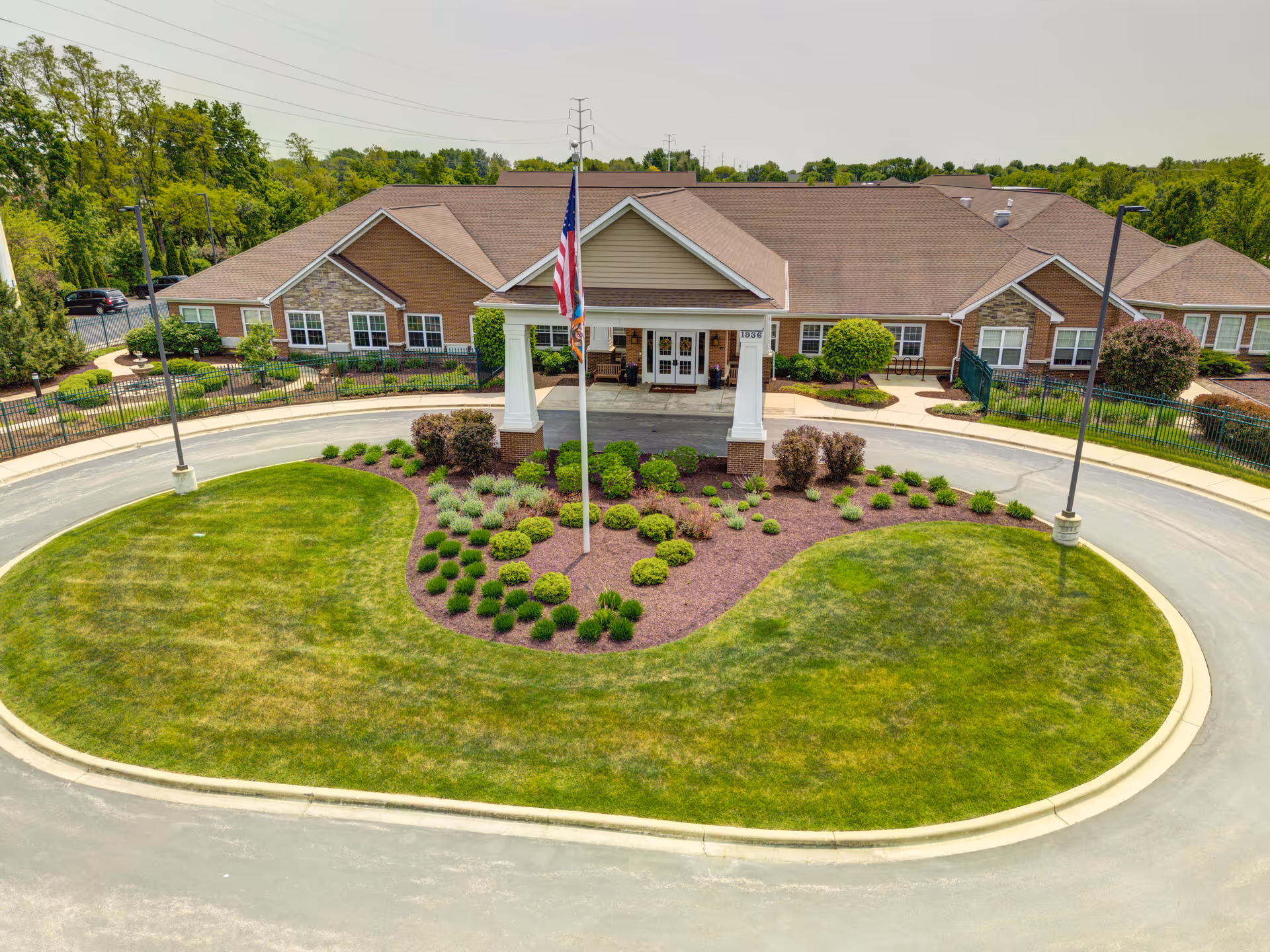Front exterior view of The Auberge at Naperville senior living facility with a circular driveway, landscaped garden with shrubs and an American flagpole in the center, and a single-story building with a covered entrance.