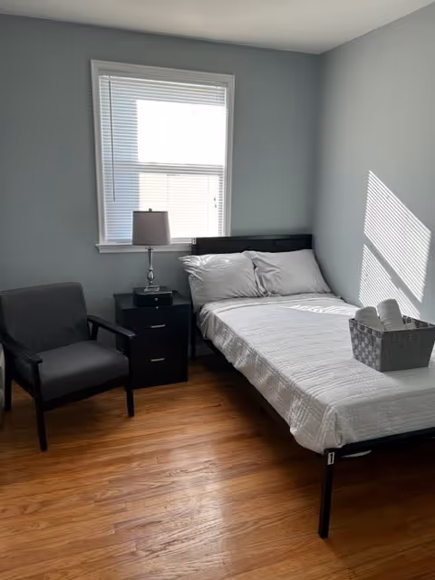 A simple furnished bedroom with a bed, nightstand, lamp, and chair by a window.
