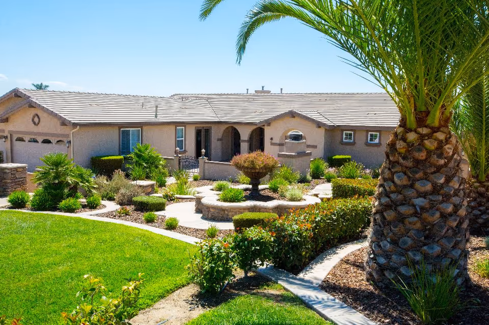 Front exterior of a single-story beige senior living building with a tiled roof, landscaped garden, walkway, and a large palm tree.