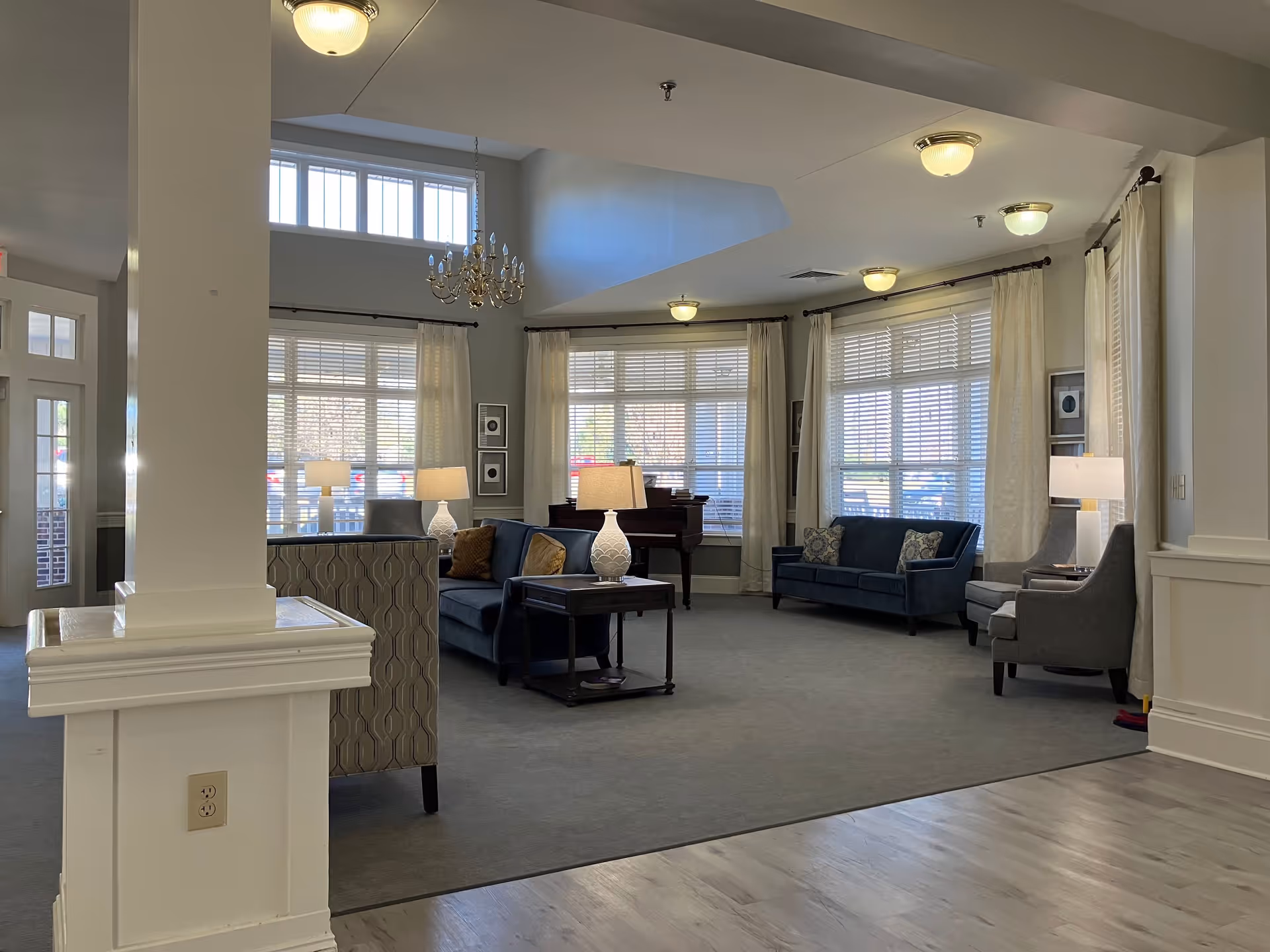 A spacious senior living facility common area with large windows covered by white blinds and cream curtains. The room features blue sofas, gray armchairs, side tables with lamps, a chandelier, and a piano in the corner. The floor is a combination of carpet and wood, and the walls are painted light gray.