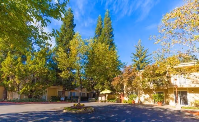 Exterior view of a facility driveway and courtyard with tall trees, a circular planted island, and a two-story building under a blue sky.