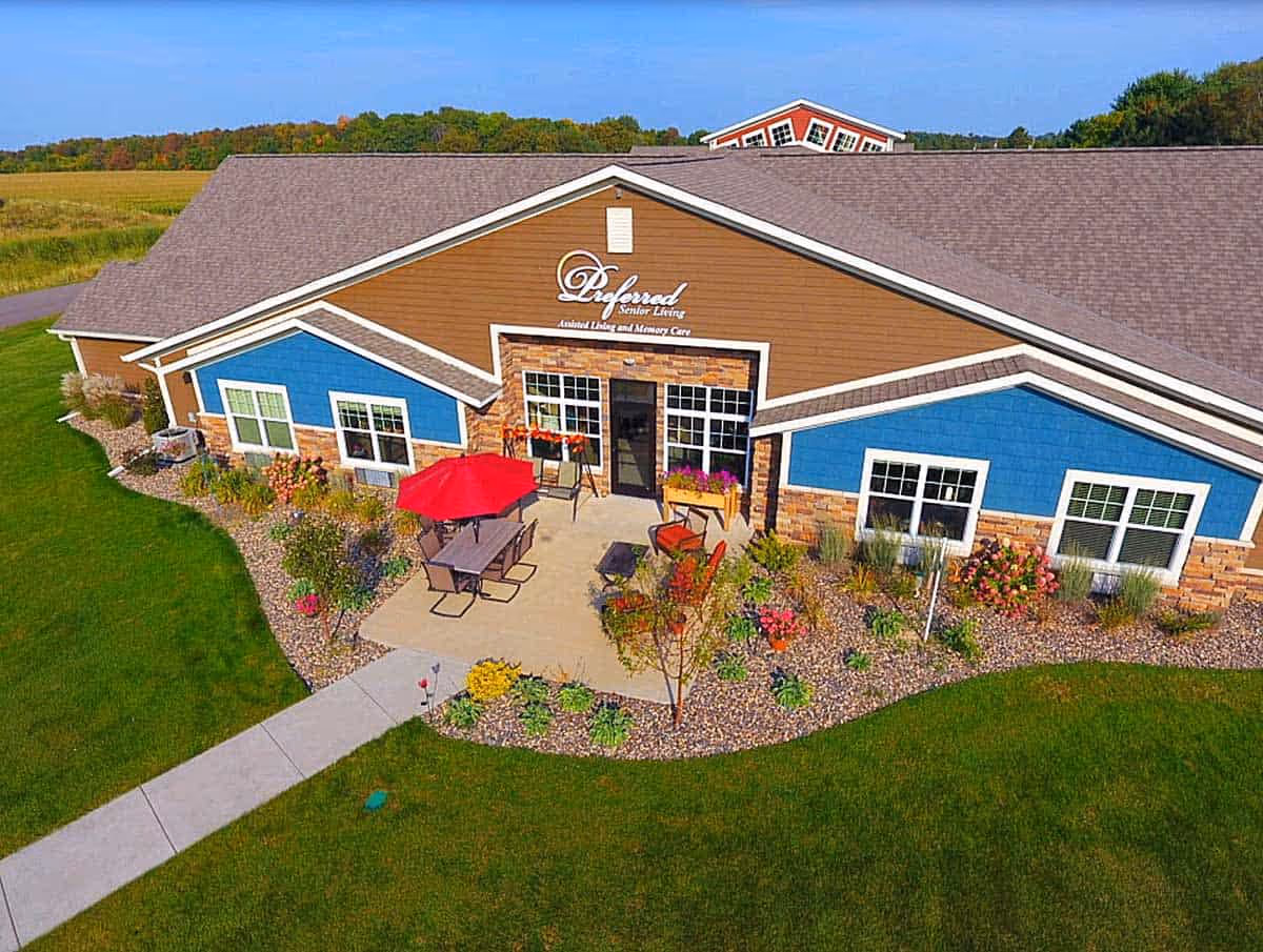 Front exterior of the Preferred Senior Living building with a patio area, red umbrella, and landscaped grounds.