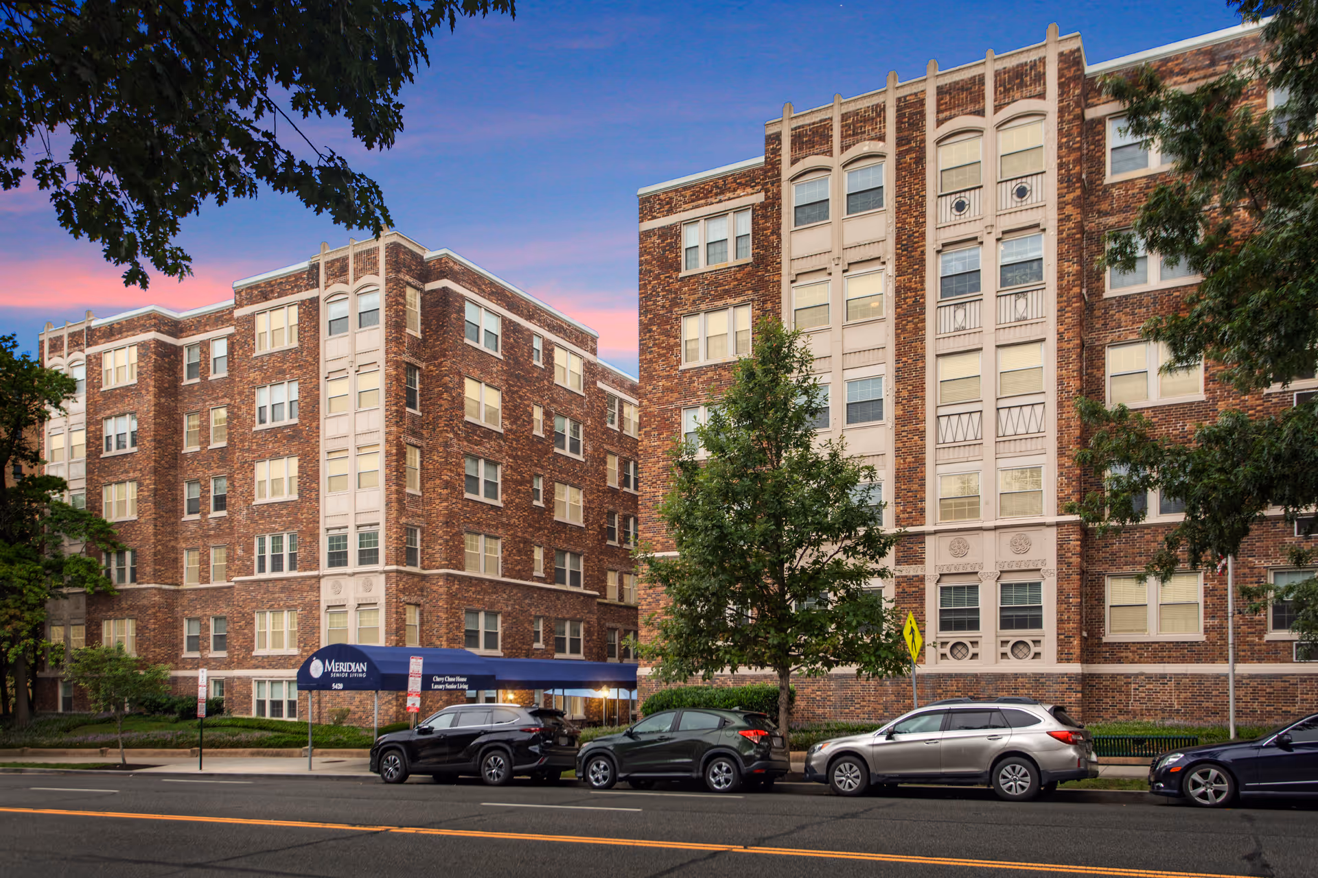 Exterior view of a multi-story brick building with decorative architectural details at dusk. Several cars are parked along the street in front of the building, which has a blue awning labeled 'Meridian Senior Living' and 'Chevy Chase House Luxury Senior Living.' Trees partially frame the scene.