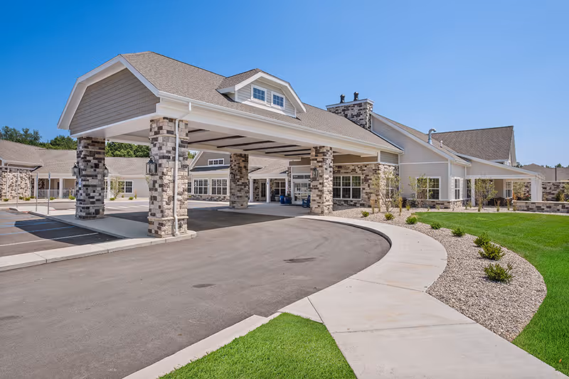 Exterior view of Quincy Place Senior Living facility showing a covered entrance with stone pillars, a curved driveway, well-maintained green lawn, and a clear blue sky.