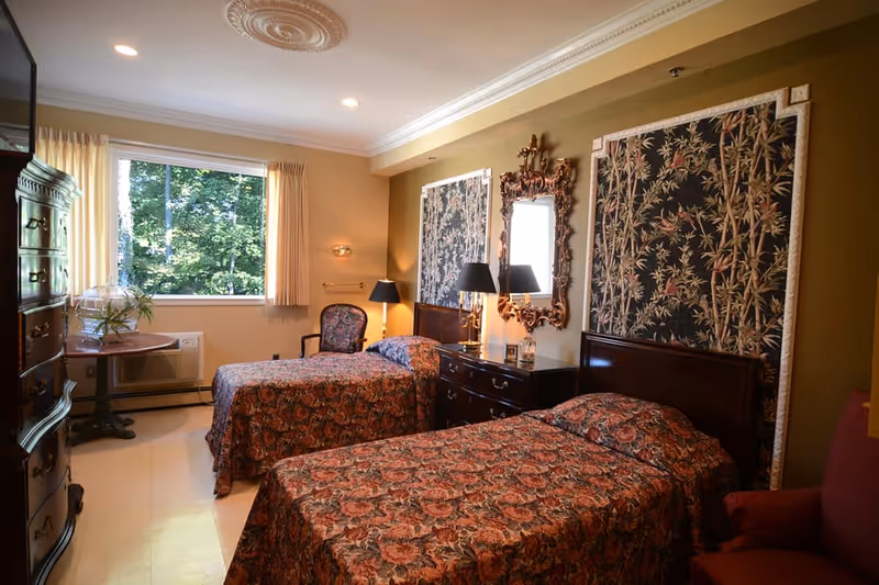 A bedroom with two twin beds covered in matching floral bedspreads. Each bed has a large decorative headboard with a floral pattern. Between the beds is a dark wooden dresser with a decorative mirror above it and two black lamps. There is a window with beige curtains letting in natural light, a wooden chair with a floral cushion, and a small round table with a glass terrarium on it. The walls are painted a warm beige color.
