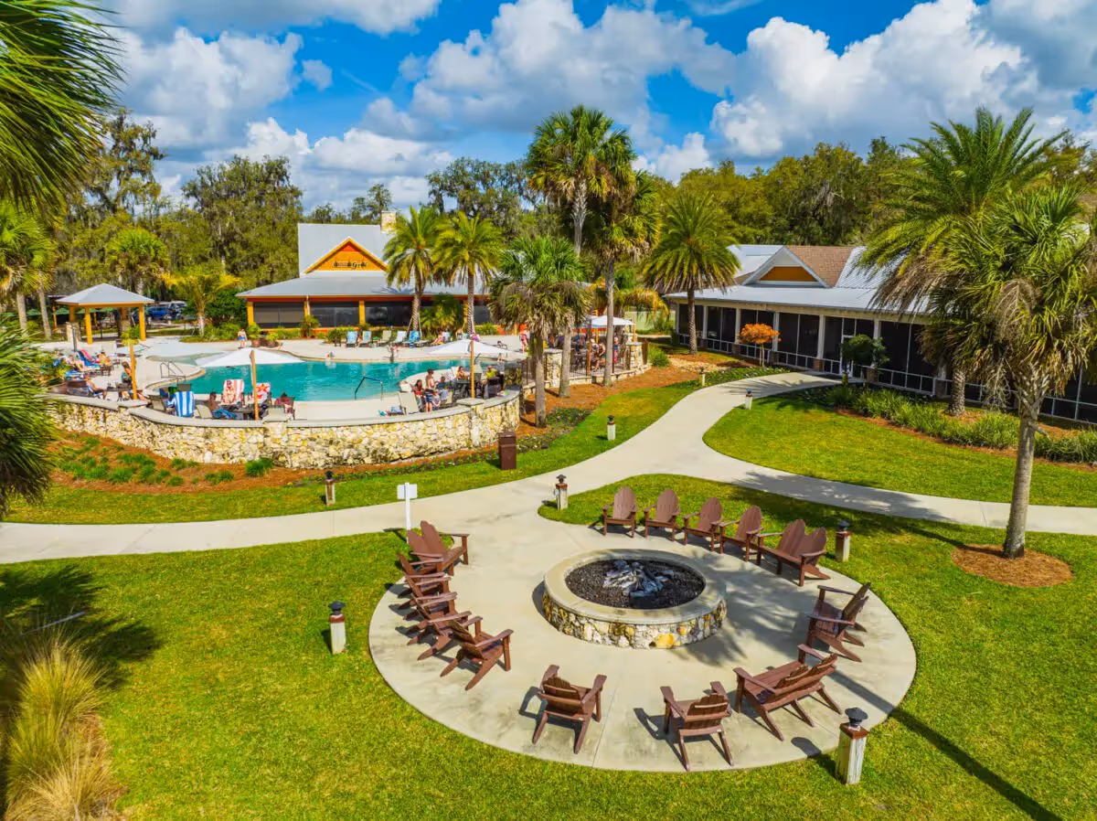 An outdoor senior living recreation area with a circular fire pit, surrounding chairs, a swimming pool, and a clubhouse building with palm trees.