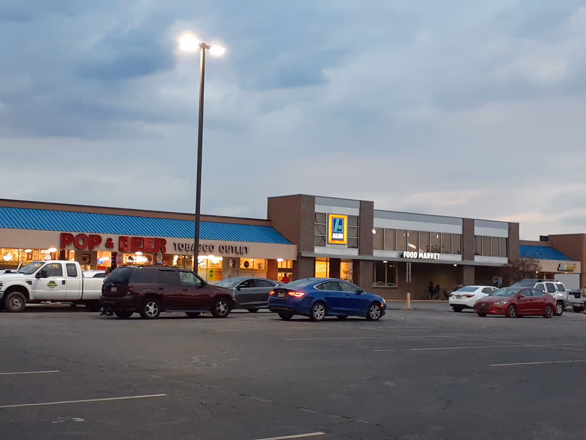 Exterior strip mall with parked cars and multiple storefronts, including a food market.