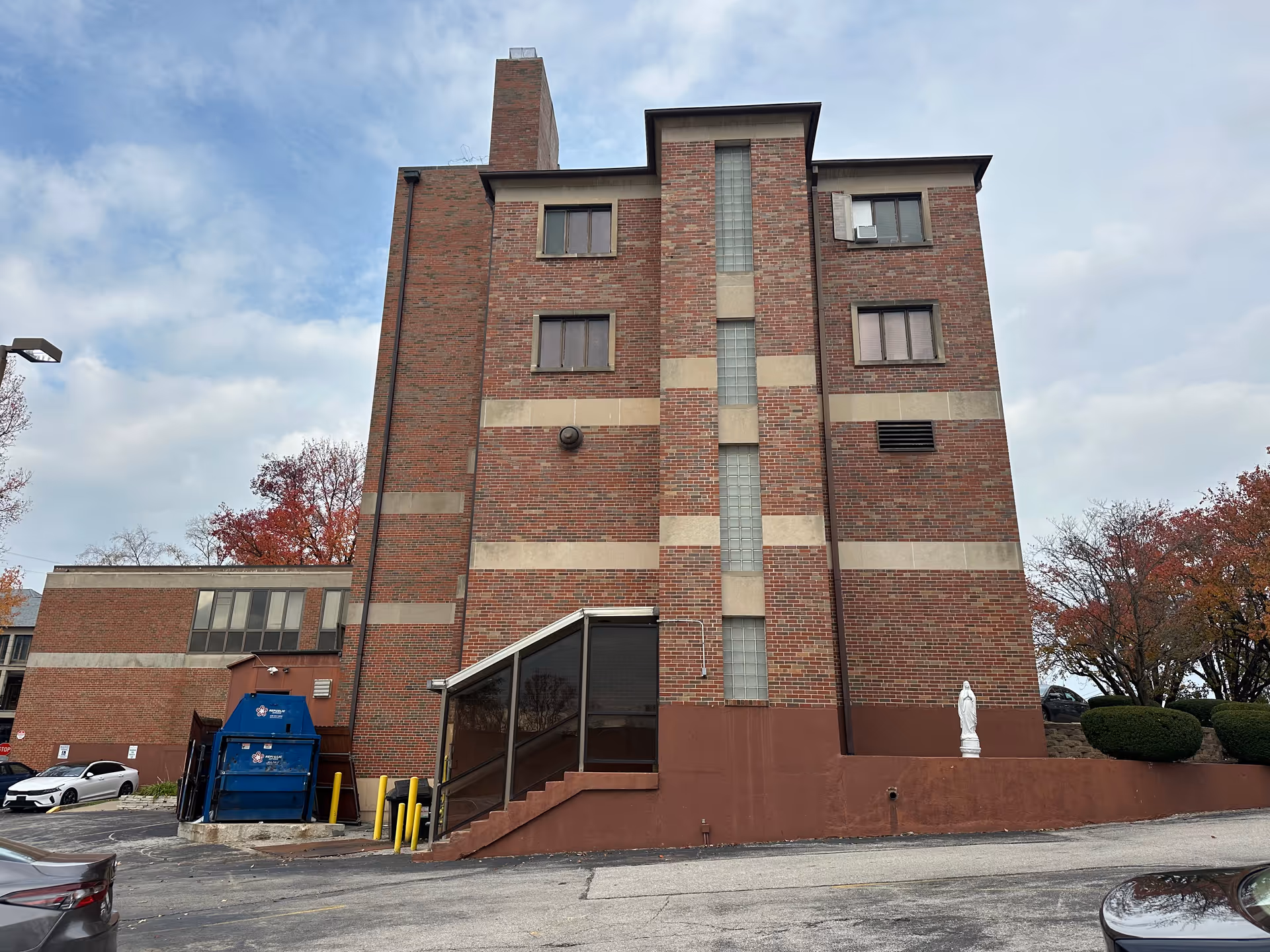 Brick four-story building with a glass-block vertical window, a small covered entrance, a blue dumpster, and a white statue in the foreground under a cloudy sky.