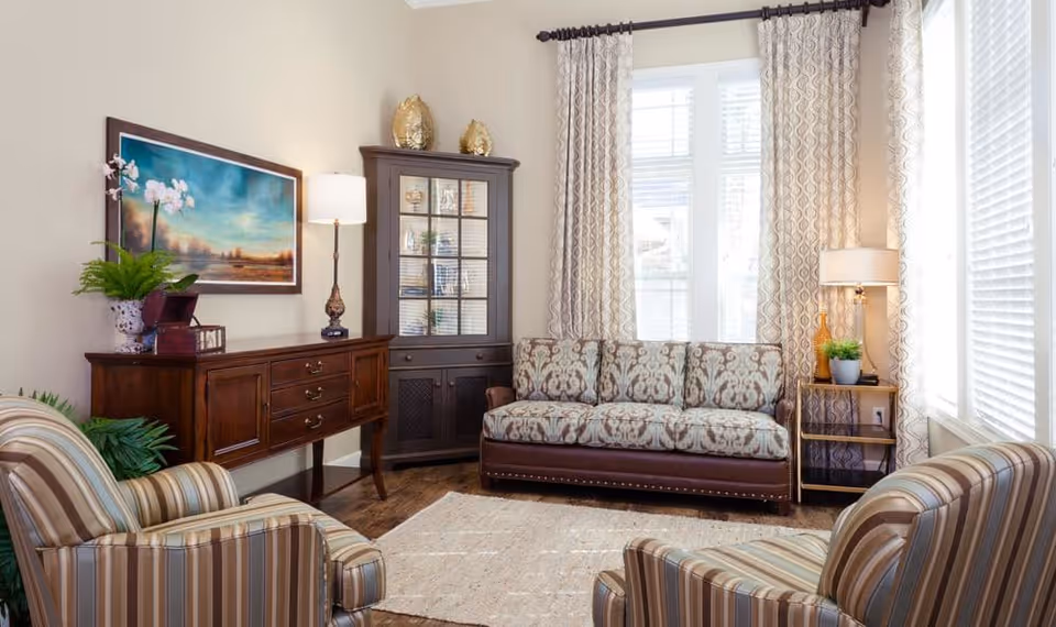 Bright, tastefully furnished living room with a patterned sofa, two striped armchairs, wooden sideboard and tall windows with curtains.