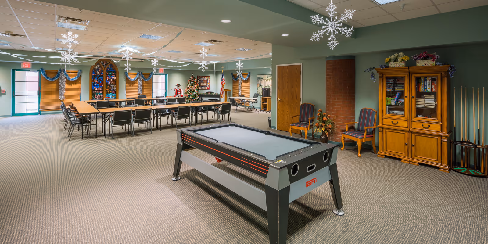 Spacious decorated community activity room with a pool table in the foreground, long tables and chairs, holiday decorations and a game cabinet.