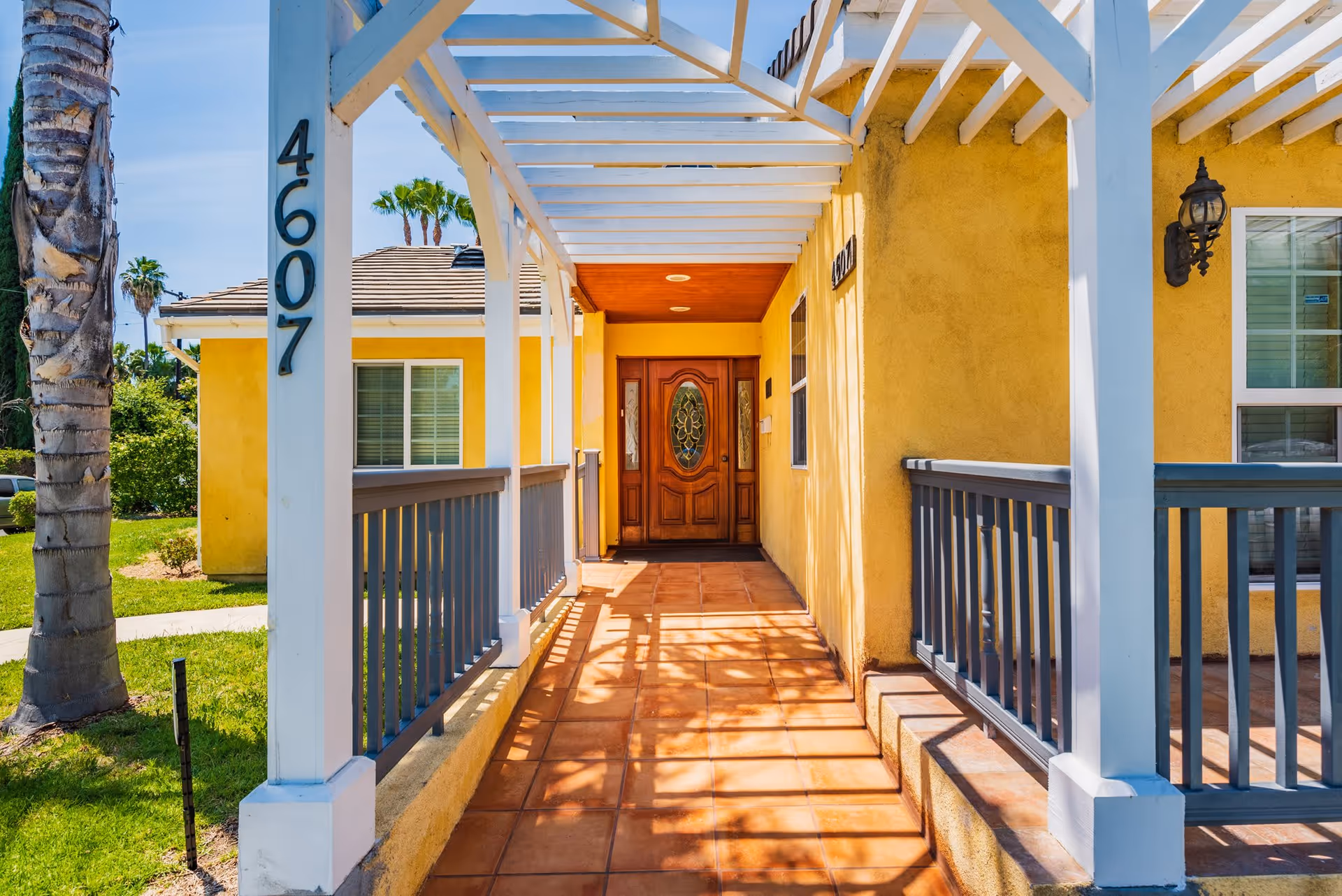 Front entrance of a yellow building with a wooden door featuring decorative glass panels. The entrance is covered by a white pergola structure with shadows cast on the tiled floor. The building number 4607 is displayed on a white post near a palm tree and green lawn.