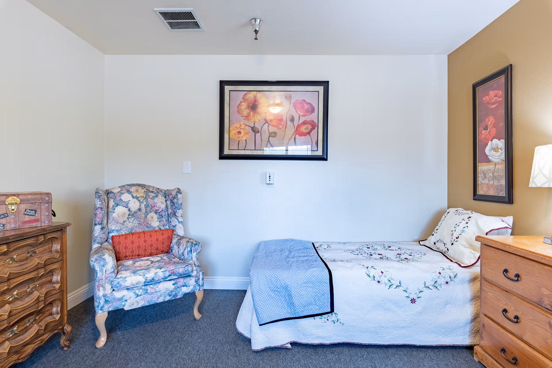 A cozy bedroom with a floral armchair, bed, and wooden dressers.