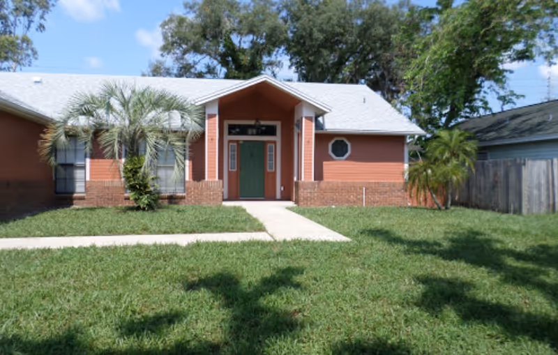 Front exterior of a single-story house with a green entry door, brick accents, a walkway, lawn, and palm trees.