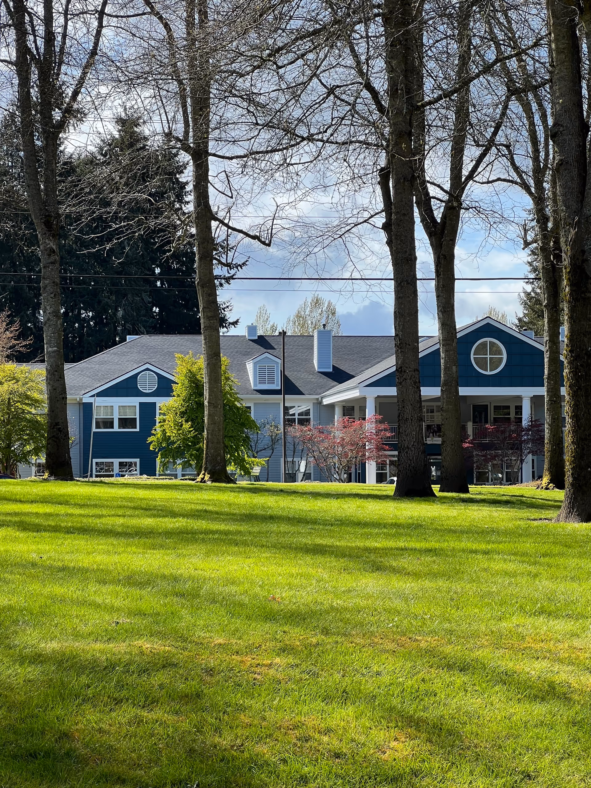 Blue assisted living building with a green lawn and tall trees in front.