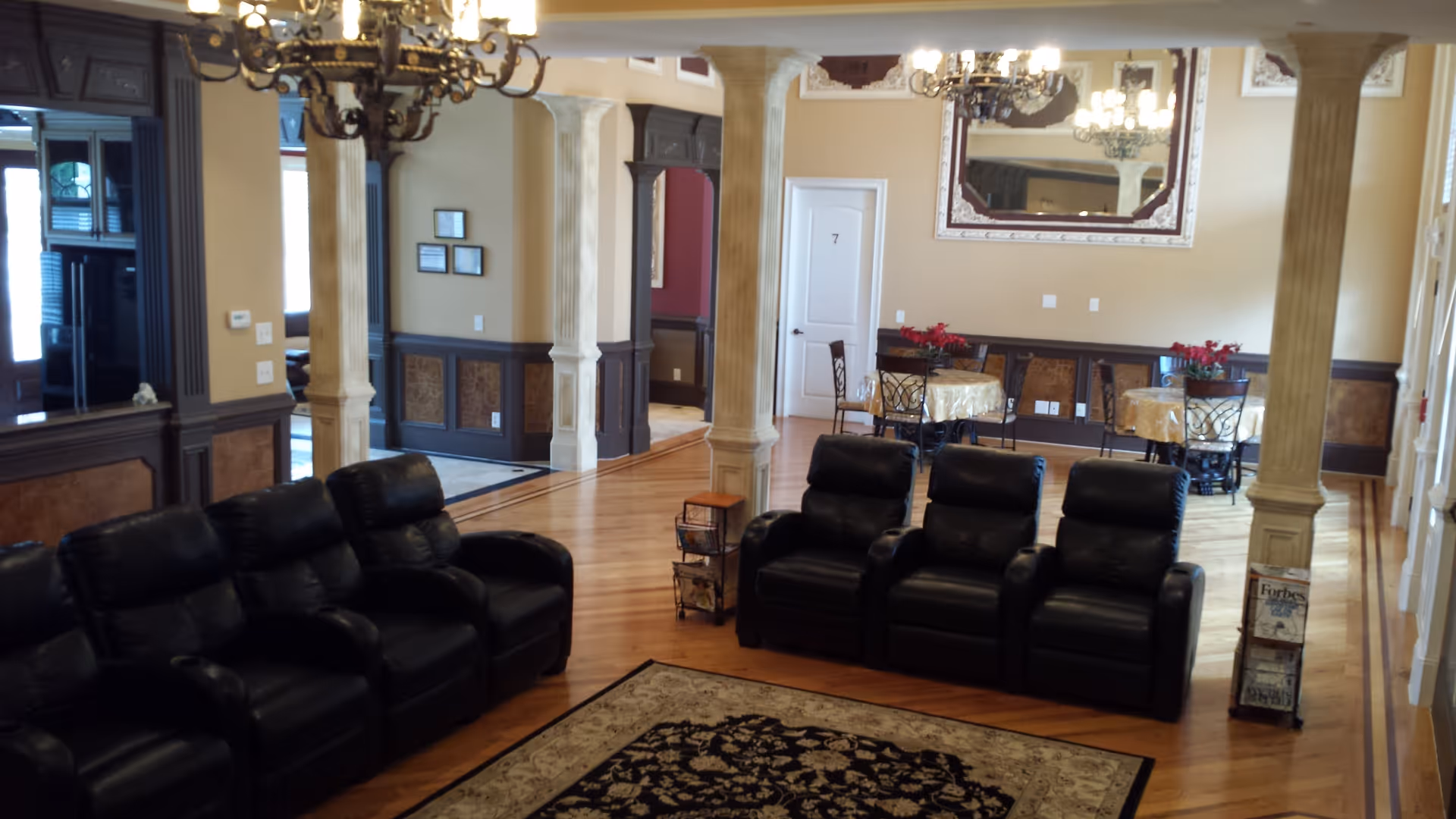 Common area with two rows of black leather recliners facing a patterned rug, decorative columns and chandeliers, and small dining tables in the background.