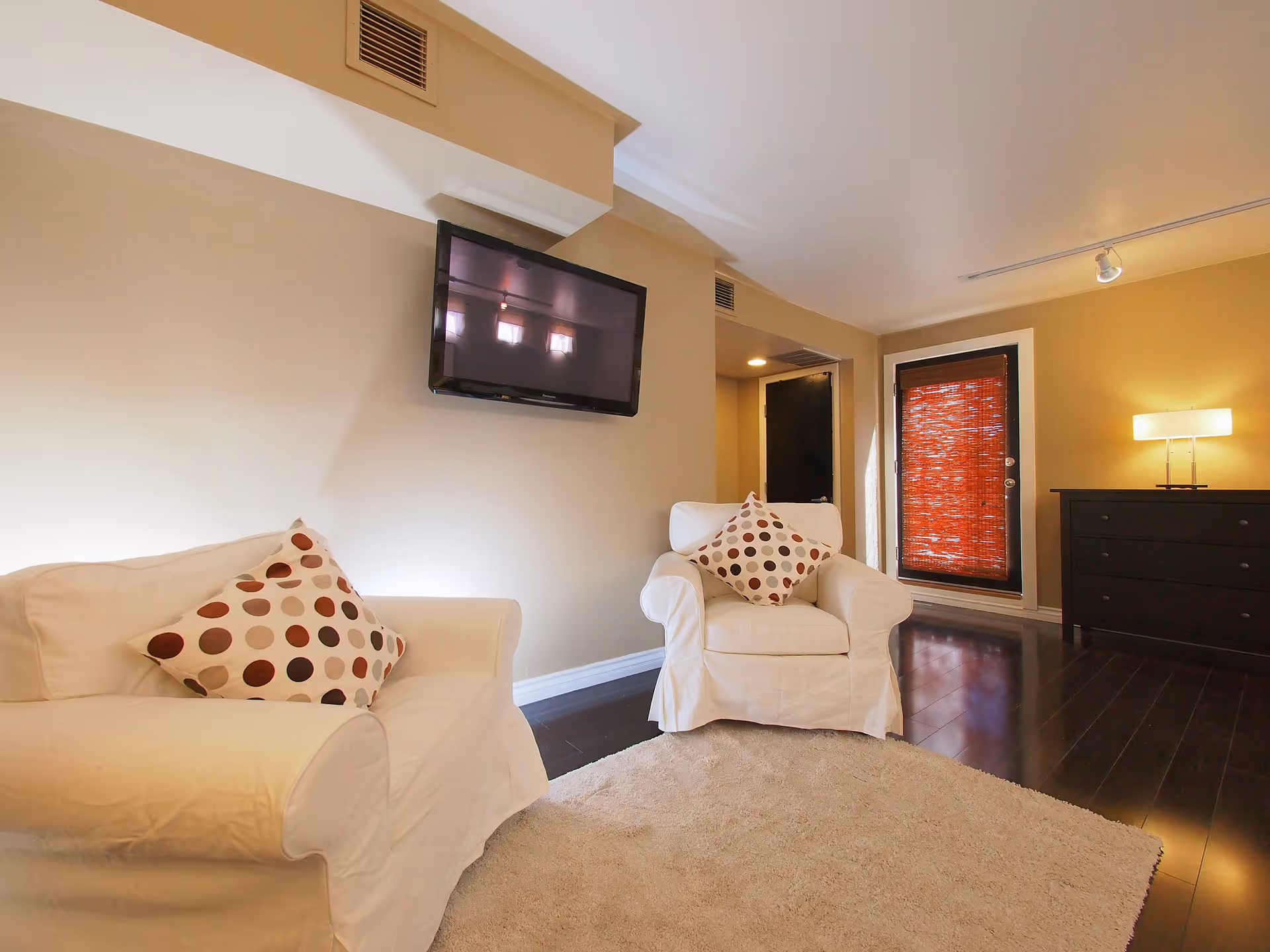 A living room with two white armchairs, a wall-mounted TV, a dresser, and a beige rug.