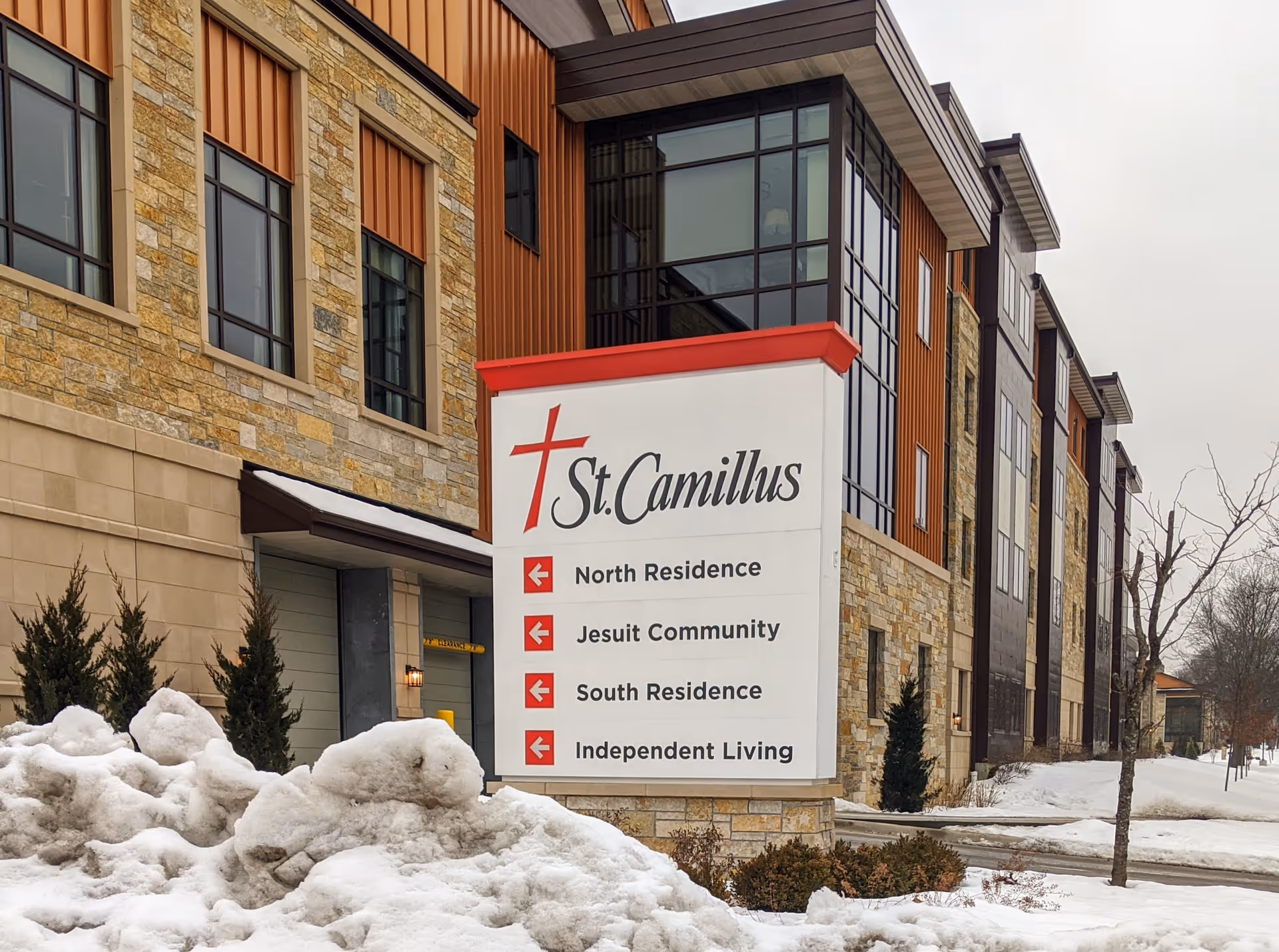 Exterior view of St Camillus Home Hospice with a directional sign amid snowy landscaping.