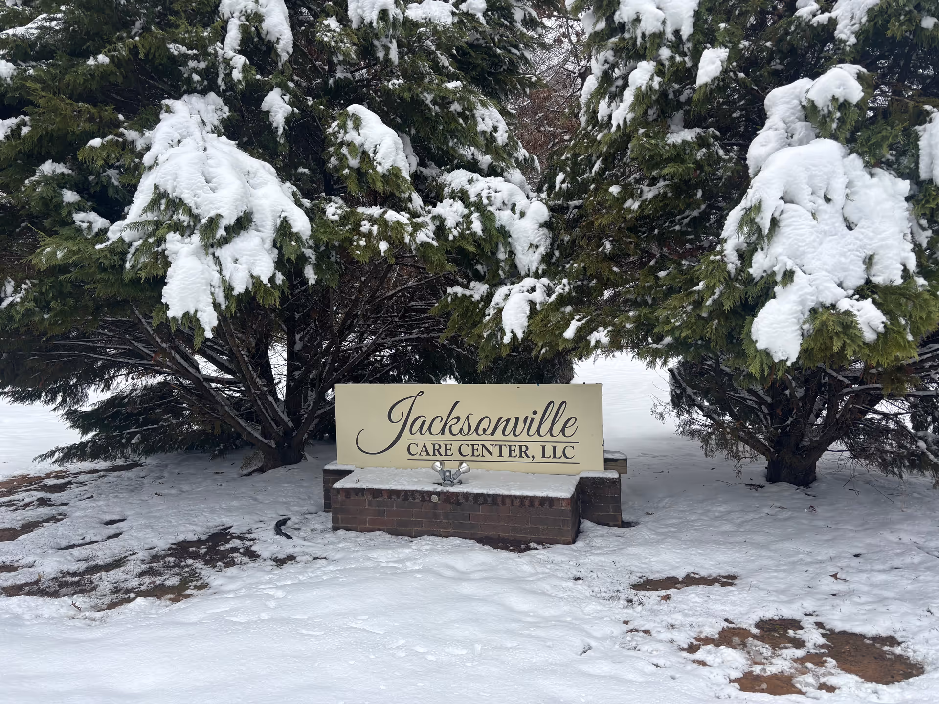 A sign for Jacksonville Care Center, LLC is placed on a brick base surrounded by snow-covered ground and evergreen trees with snow on their branches.