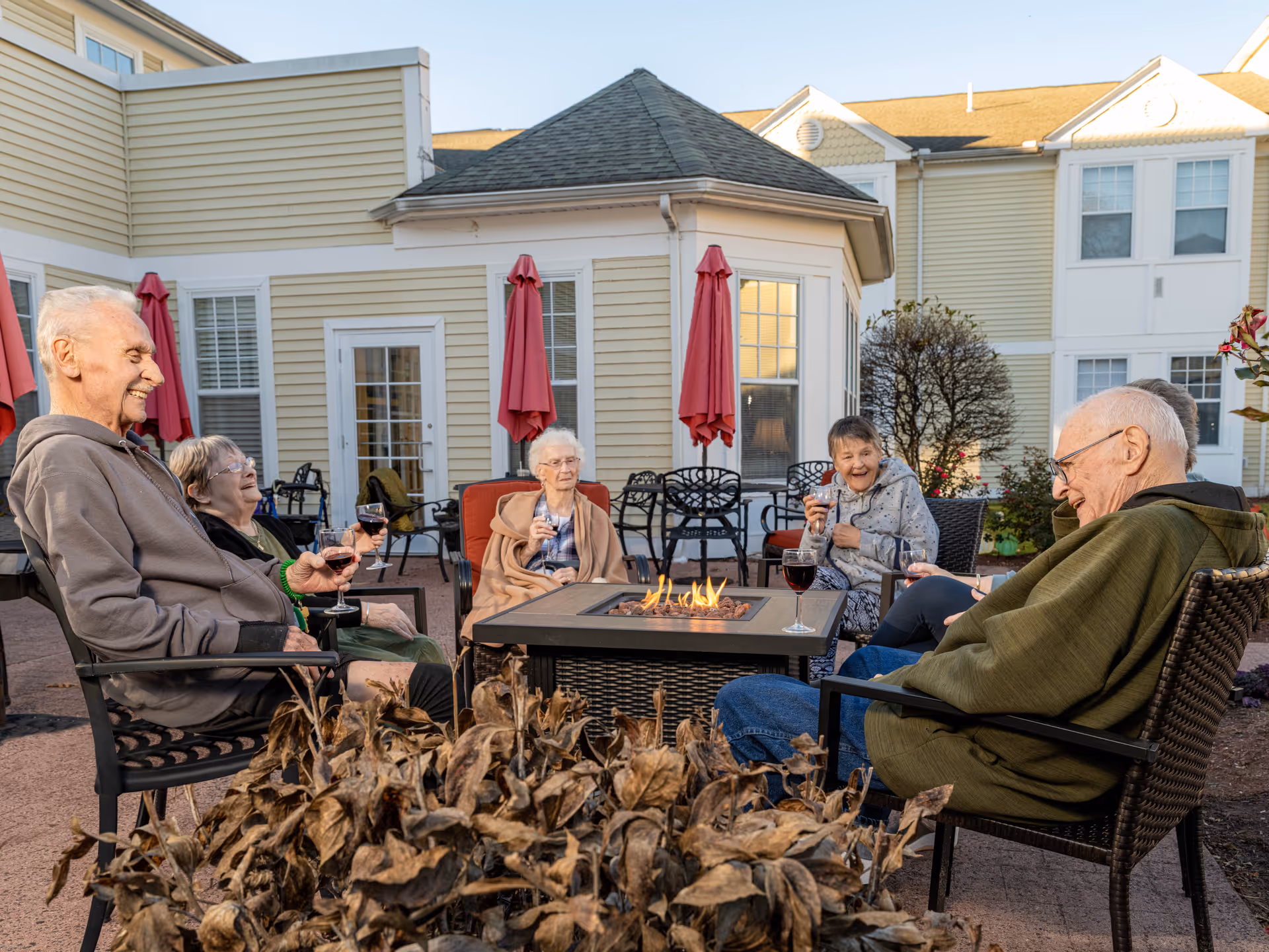 A group of elderly residents sit around a patio fire pit enjoying drinks outside a yellow assisted living building.