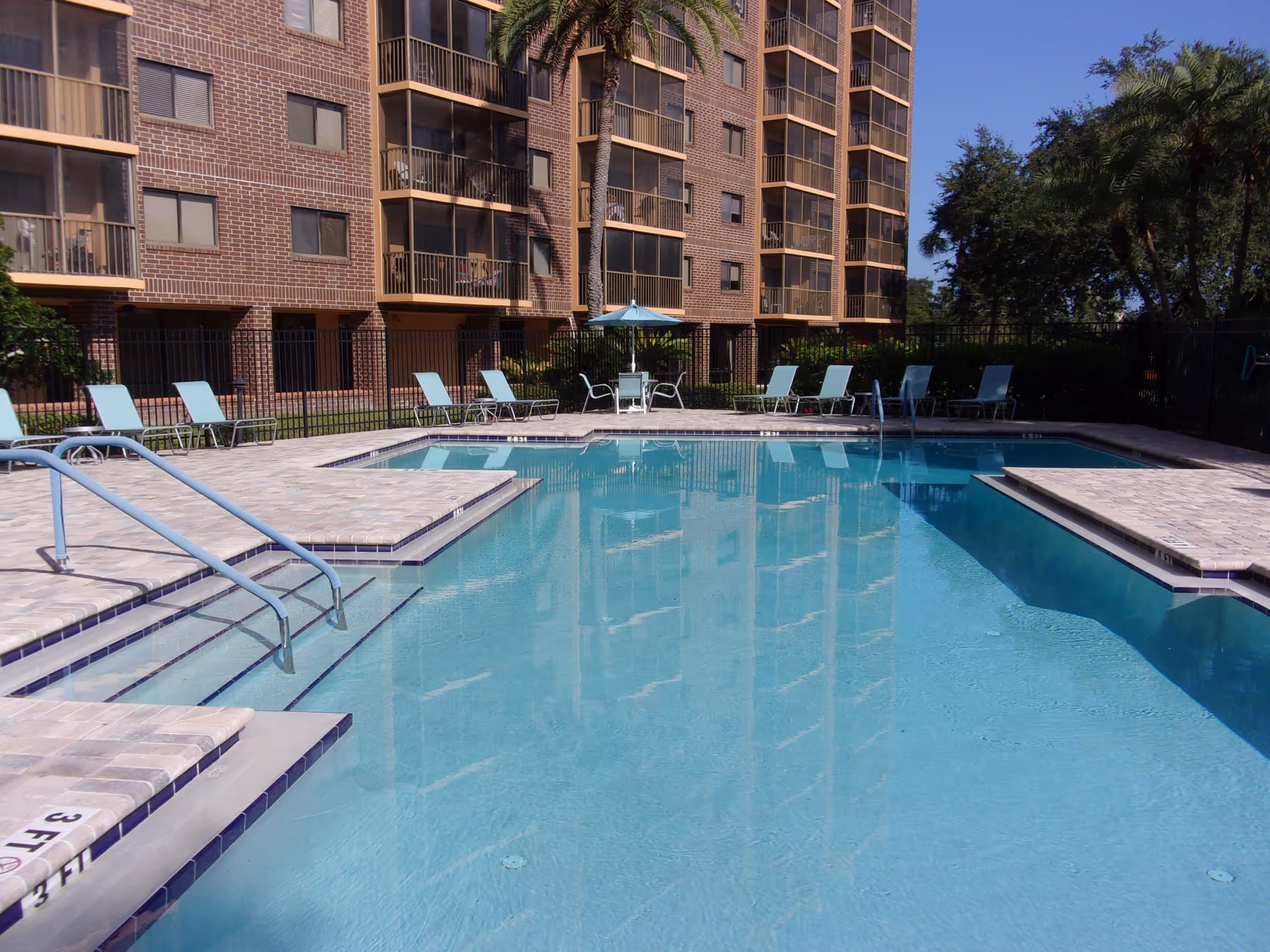 Outdoor swimming pool with clear blue water, surrounded by a tiled deck with several light blue lounge chairs and a table with an umbrella. In the background, there is a multi-story brick apartment building with balconies and windows, and some trees under a clear blue sky.