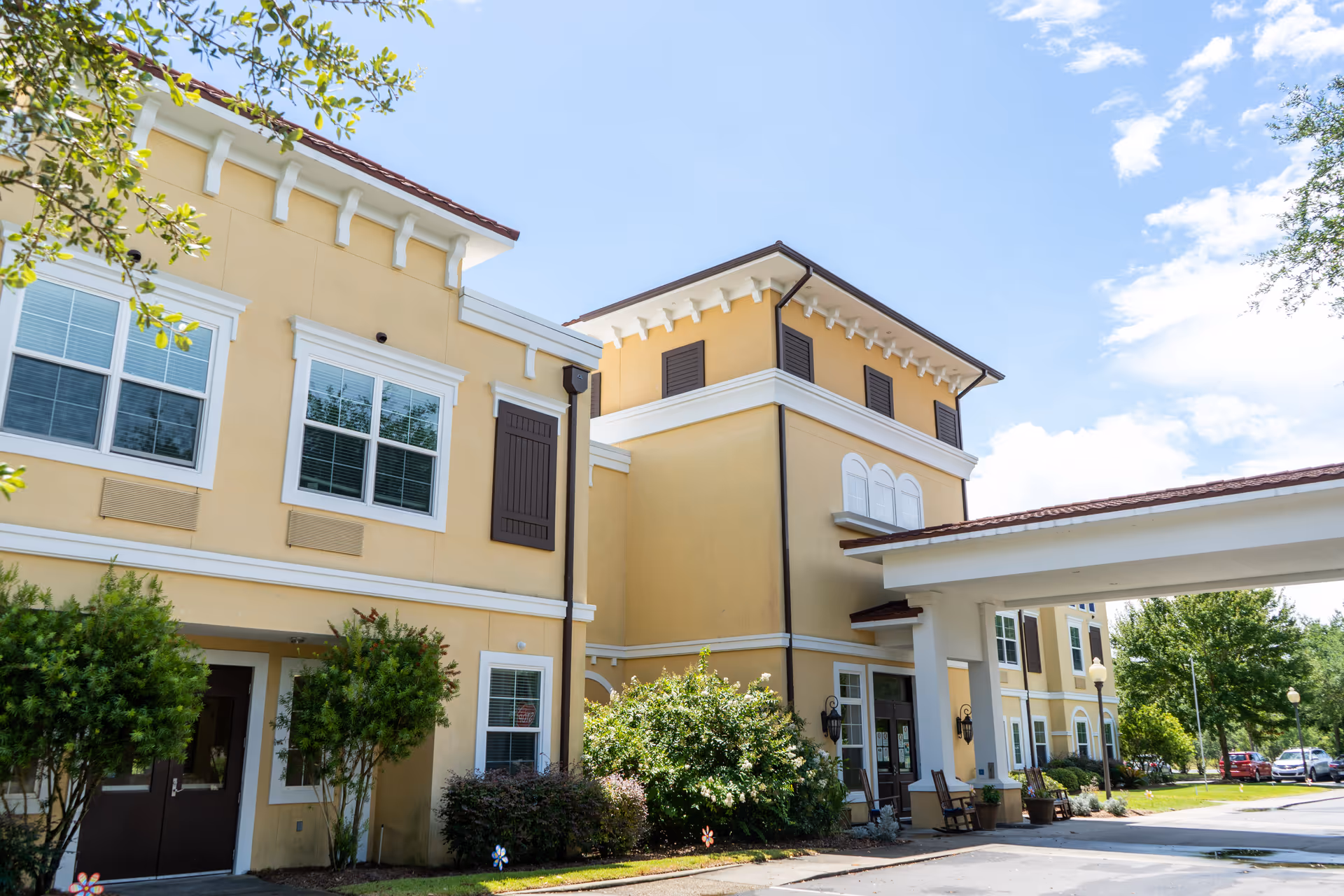 Exterior view of a senior living facility building with yellow walls, white trim, and brown shutters under a blue sky with some clouds. There are bushes and trees around the entrance, which has a covered driveway with rocking chairs nearby.