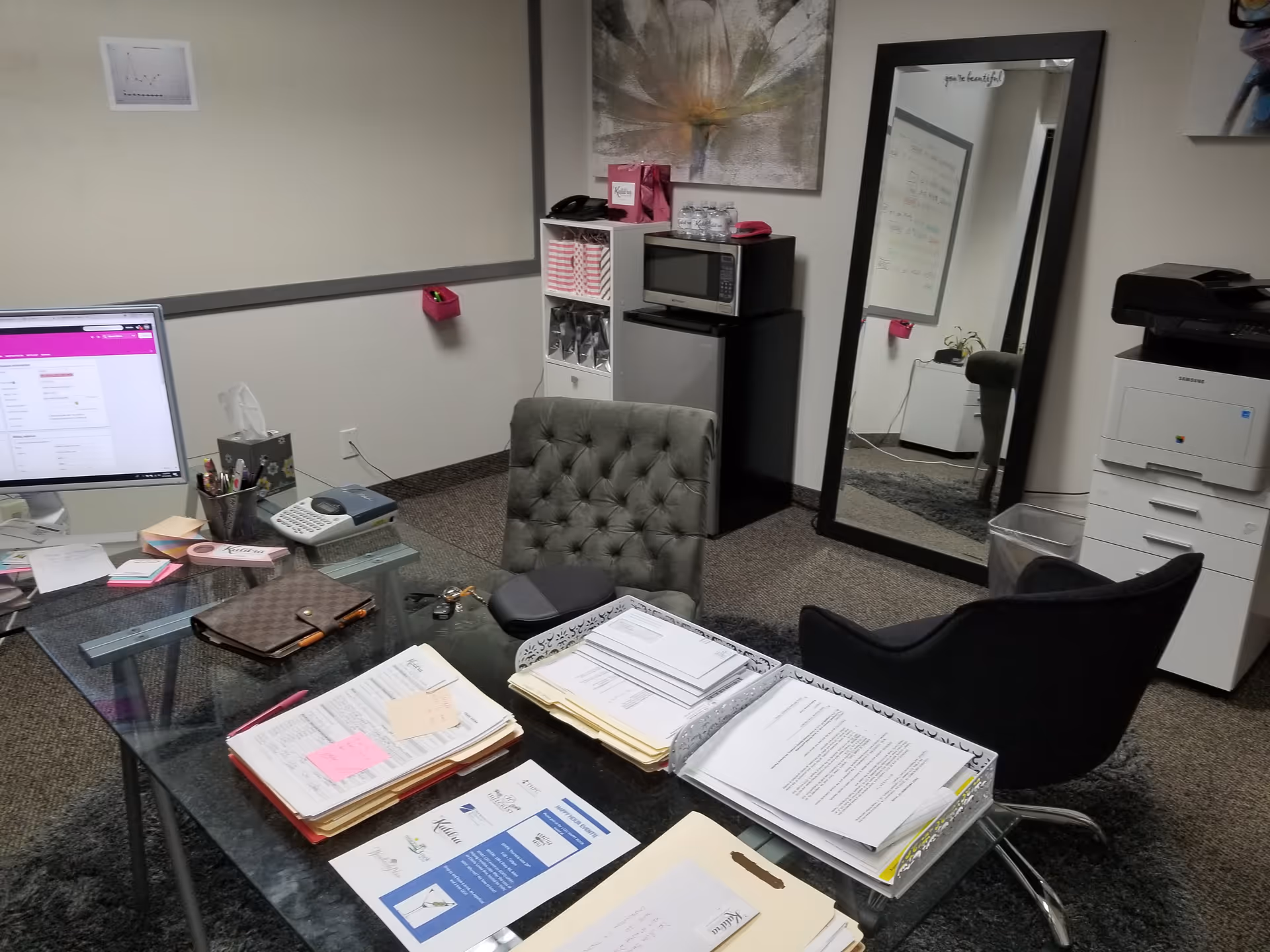 A cluttered office workspace with glass desk, computer, paperwork, and a mirror in a modern room.