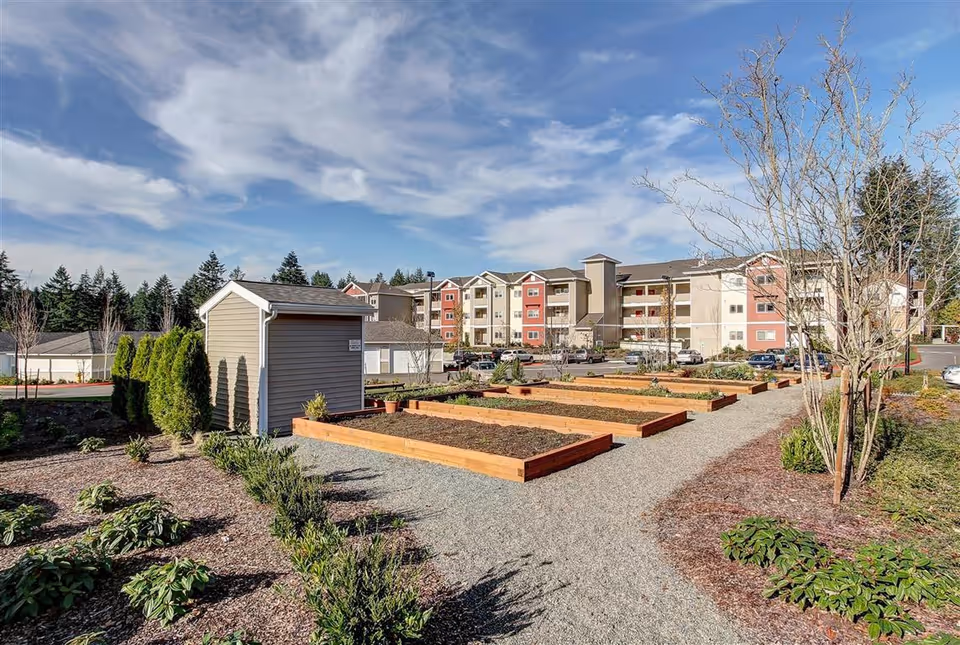 Outdoor garden area with raised wooden planting beds and a small shed, surrounded by landscaped plants and trees, with a multi-story residential building in the background under a partly cloudy sky.